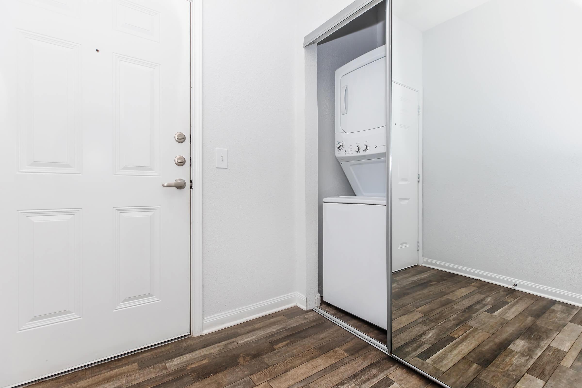 A well-lit entryway featuring a white front door and a mirrored closet door. Inside the closet, a stacked washer and dryer are visible. The flooring is made of dark wood, complementing the light walls. The space appears clean and organized, creating an inviting atmosphere.