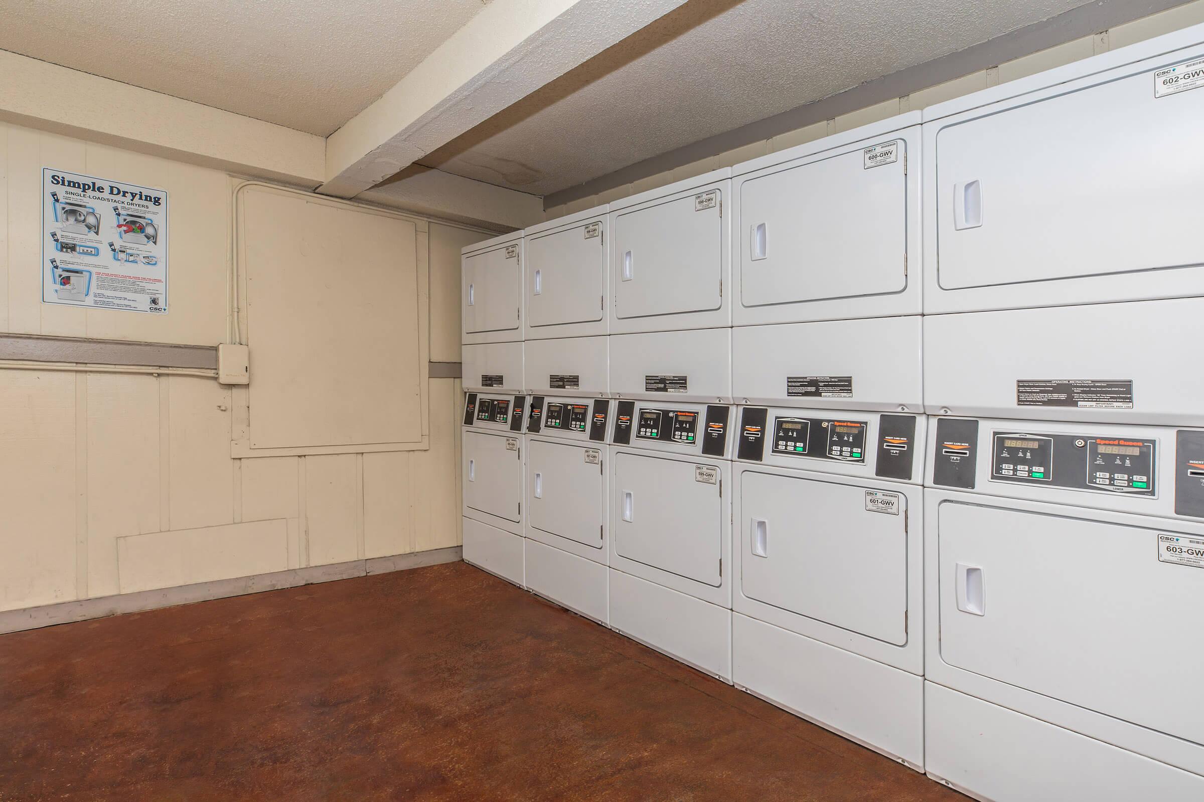 A laundry room featuring multiple rows of white dryers. Each dryer has a control panel with buttons and a digital display. The room has a clean, well-lit appearance, with a poster on the wall providing information about the drying services. The flooring is a warm, reddish-brown color.