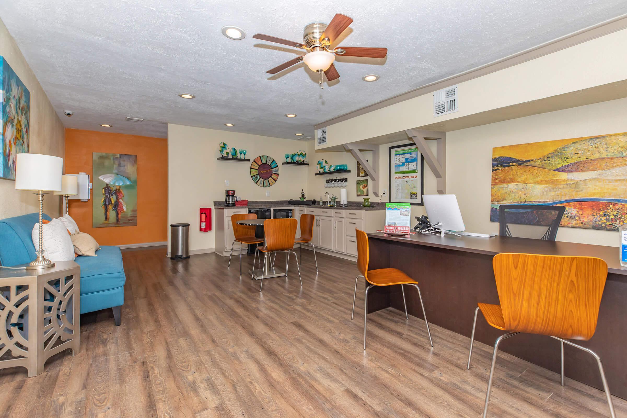A cozy reception area with a blue couch and wooden chairs, featuring a brightly colored wall with artwork. The front desk has a modern design, and there is a small kitchen or refreshment area in the background. The space is well-lit and inviting, with a ceiling fan and decorative elements.