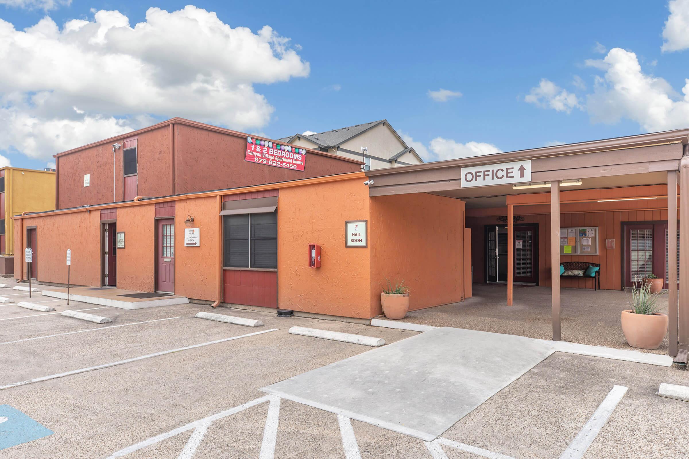 Exterior view of a colorful commercial building with orange and brown siding. The building features an "OFFICE" sign with an arrow pointing towards the entrance. There are multiple doors, a parking lot in front, and a sign indicating an office. The sky is bright with scattered clouds.