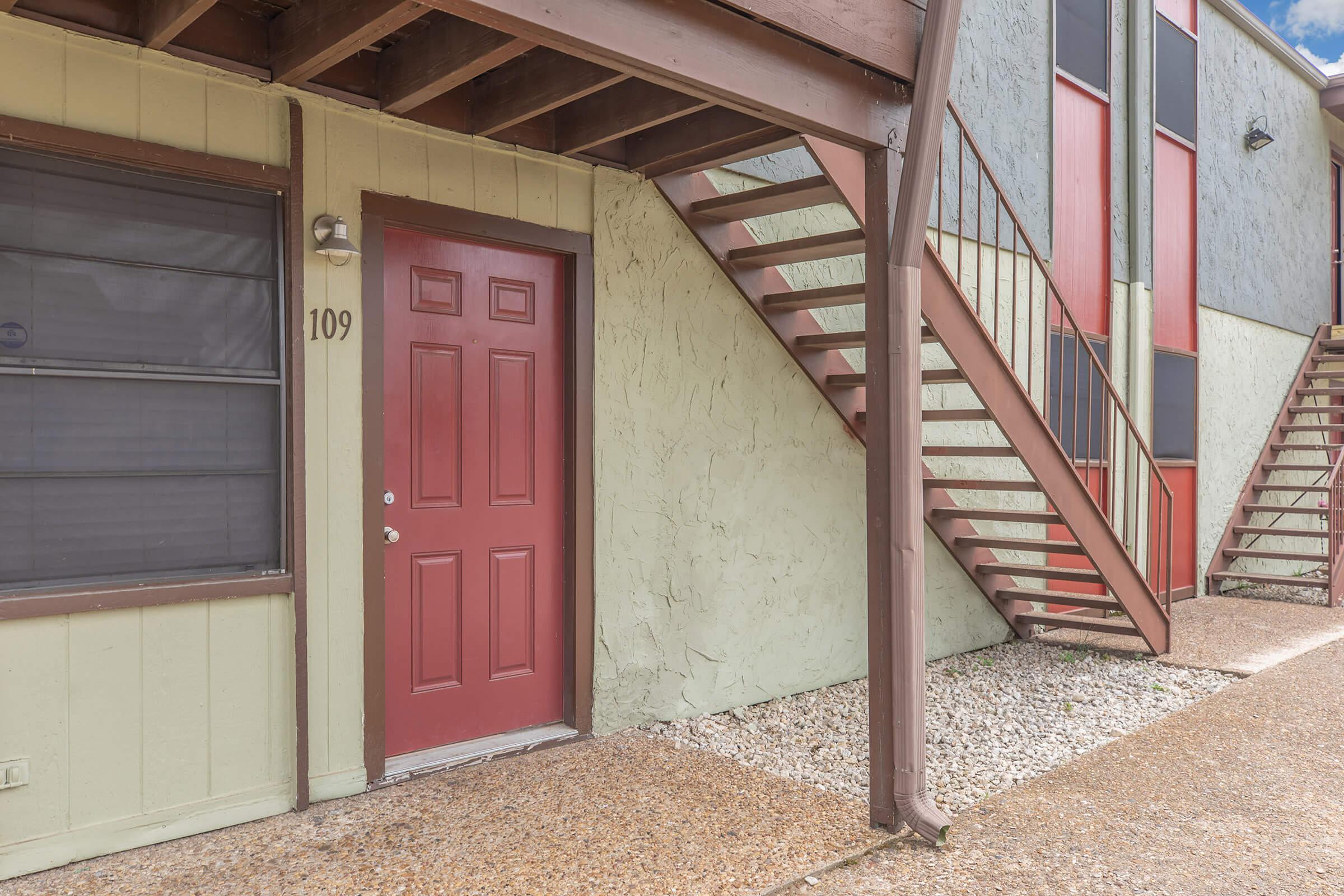 Exterior view of an apartment building featuring a red door labeled "109." The entrance is flanked by a staircase leading to the upper level. The building has a light green exterior with a textured finish and a gravel walkway in front. The bright blue sky is visible above.