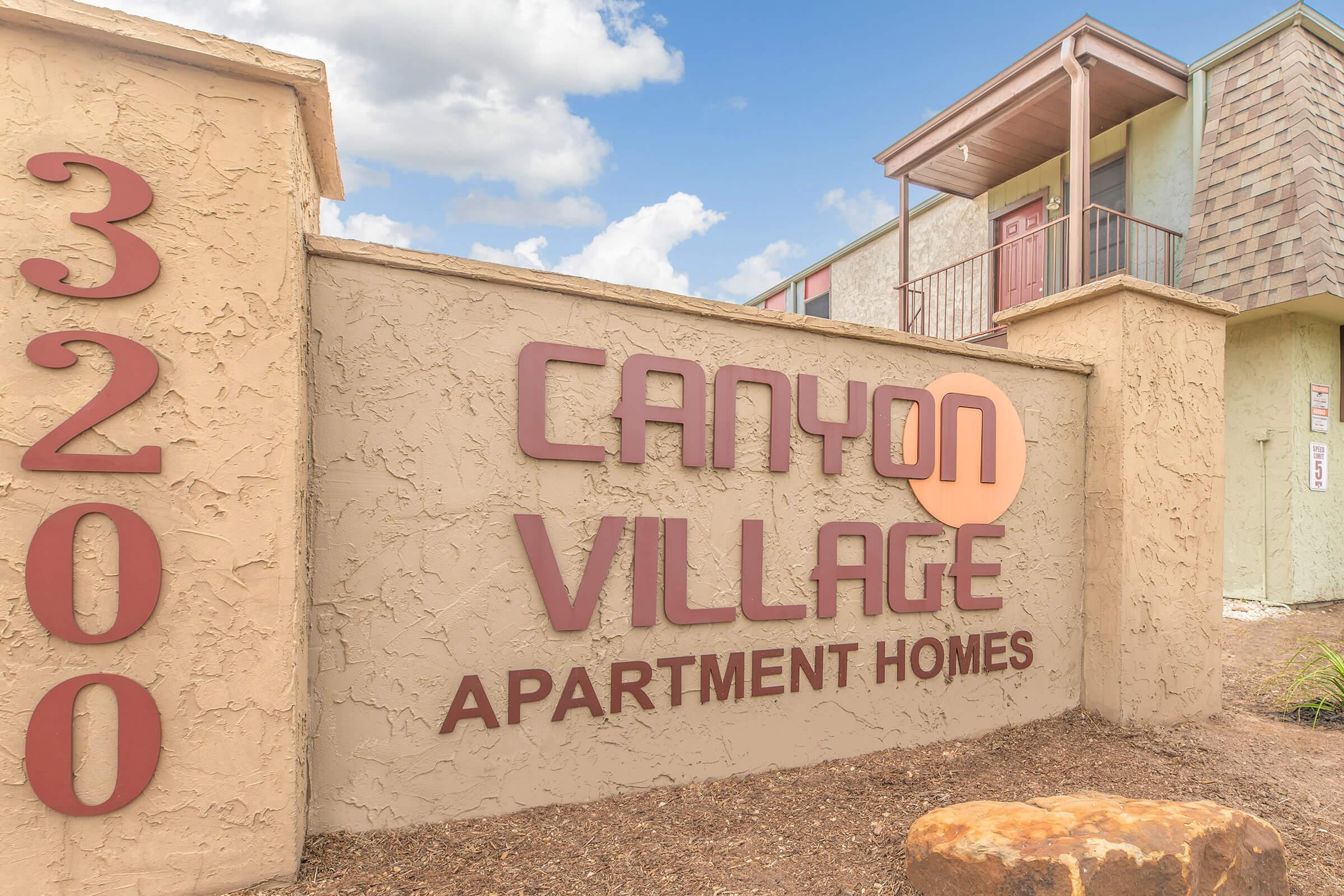 Sign for Canyon Village Apartment Homes located at 3200, featuring a modern design with a light color palette. The sign highlights the name "Canyon Village" in large letters, indicating a residential community. The background shows a blue sky with clouds.