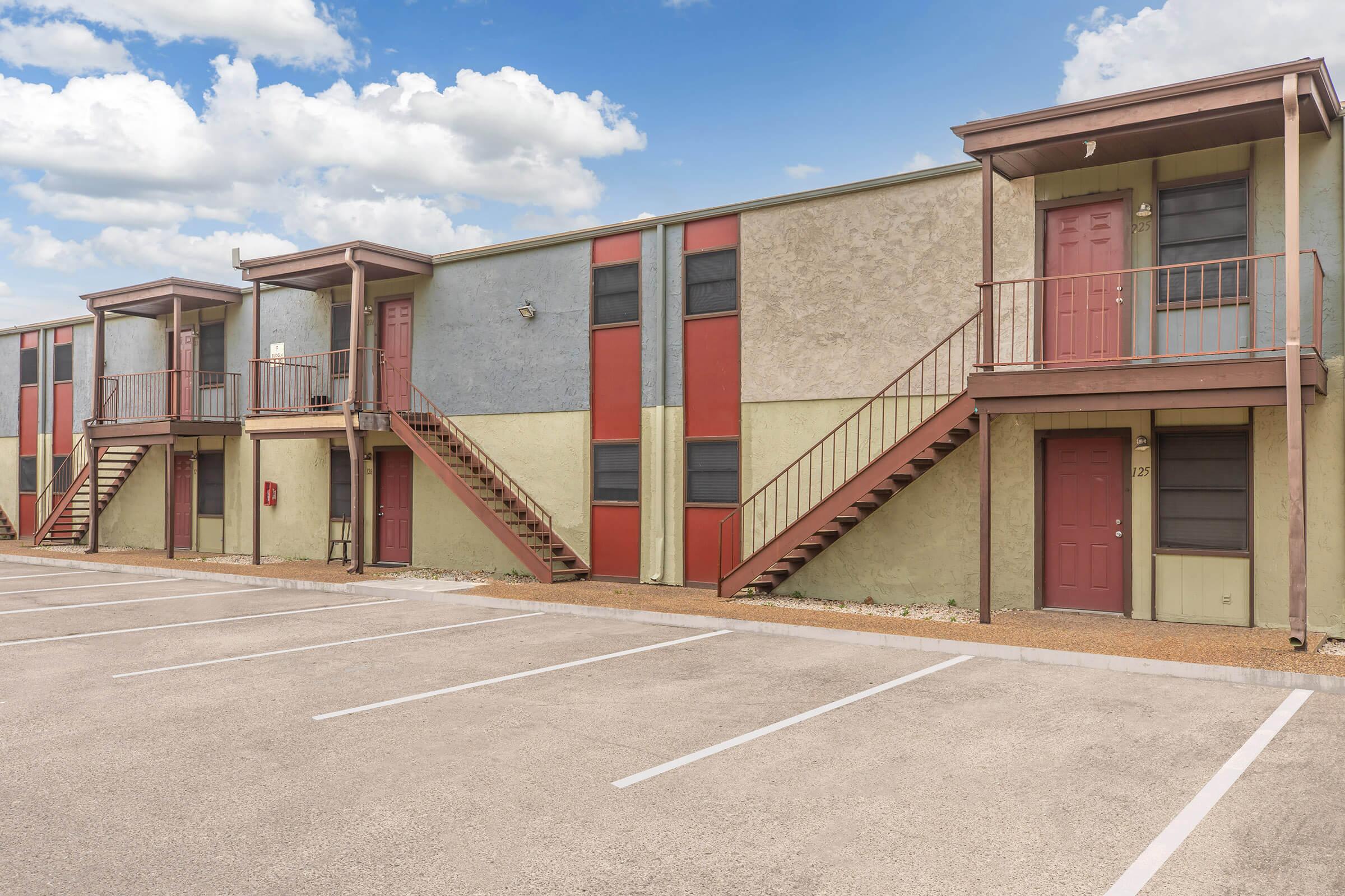 Multicolored apartment building with two levels, featuring red and blue walls, and staircases leading to entrances on both sides. A parking lot is visible in front, with marked parking spaces and a gravel area beneath the building. The sky is partly cloudy.