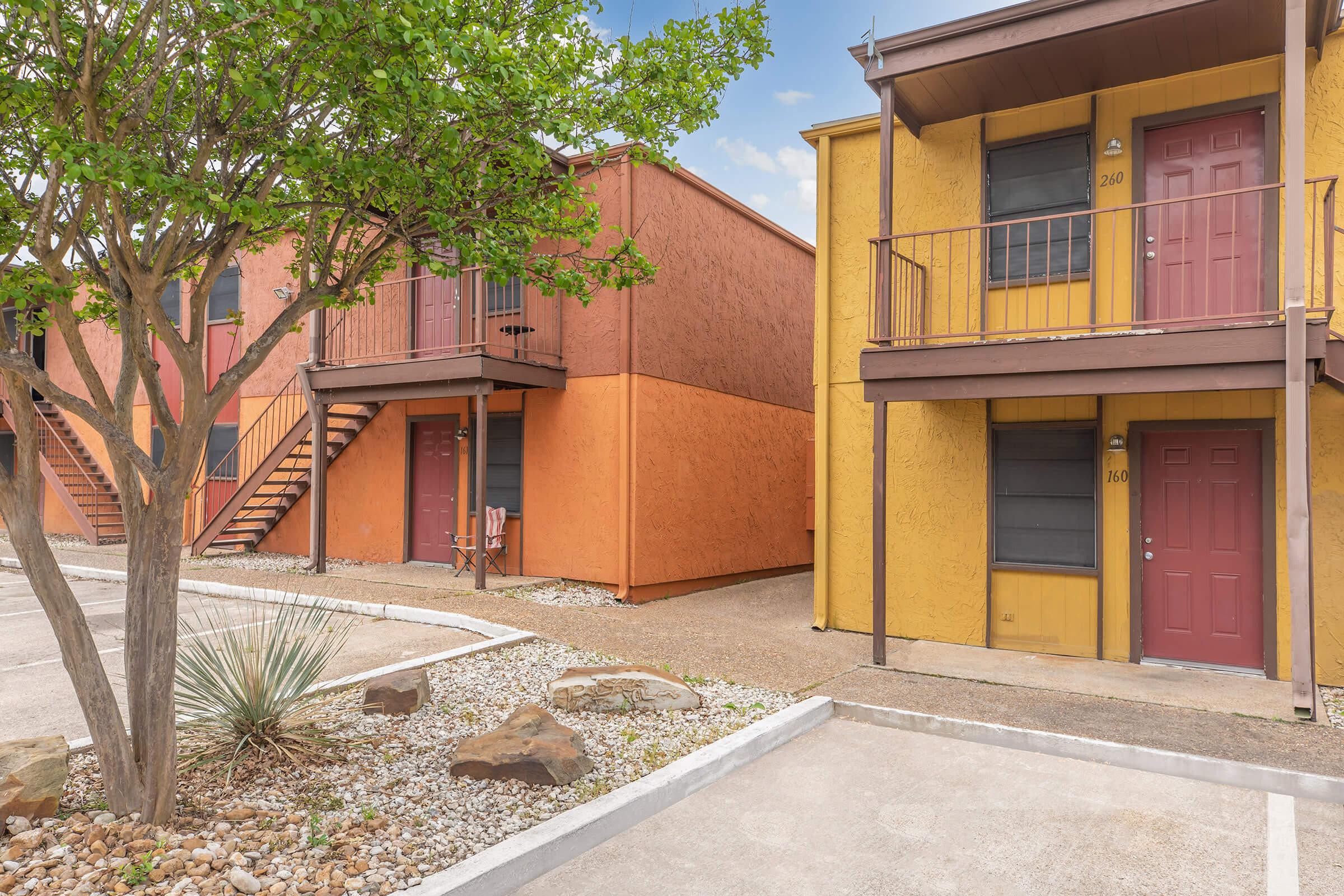 A view of a courtyard featuring two colorful apartment buildings, one in orange and the other in yellow. Each building has a staircase, balconies, and entrances with red doors. The area is landscaped with small rocks and a tree, set against a clear blue sky.
