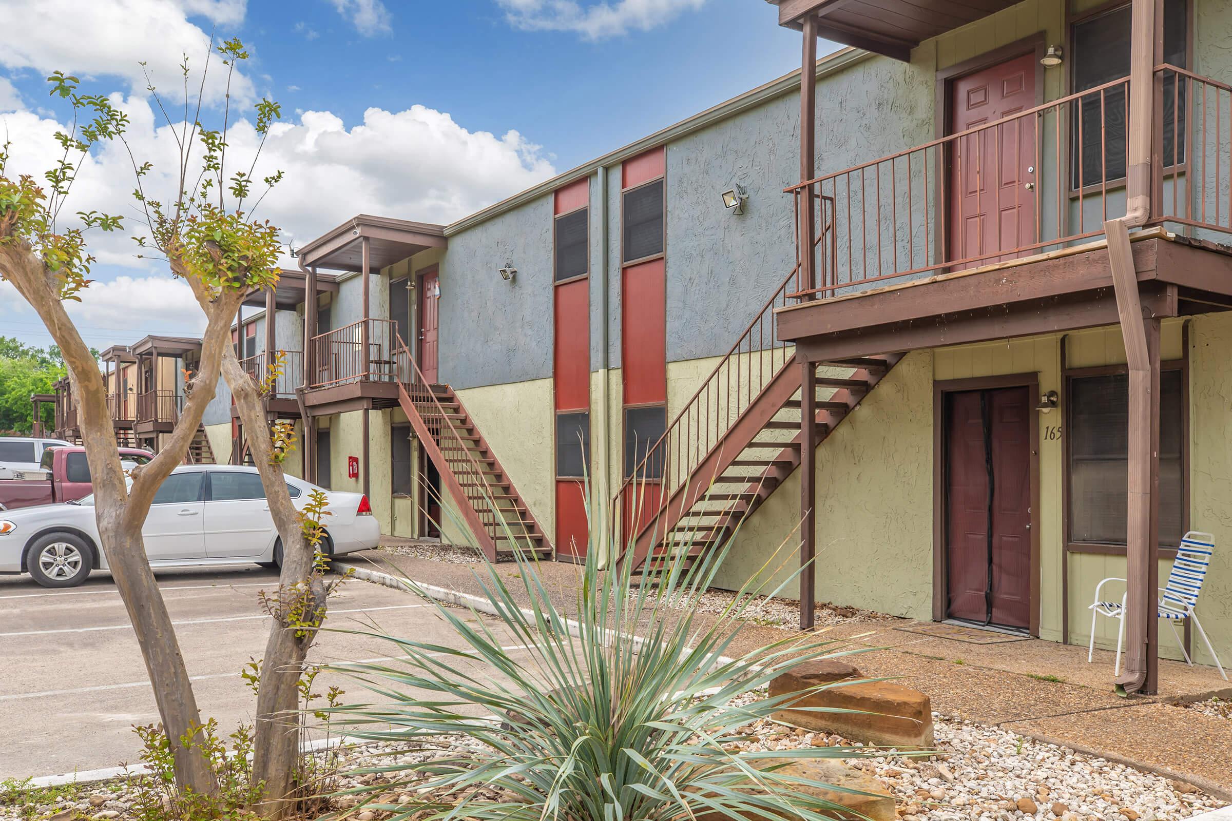 A view of a two-story apartment complex featuring several units with colorful exteriors in shades of green and red. Stairs lead to the second floor, and a small landscaped area with a tree and ornamental plants is visible in the foreground. A white car is parked nearby.