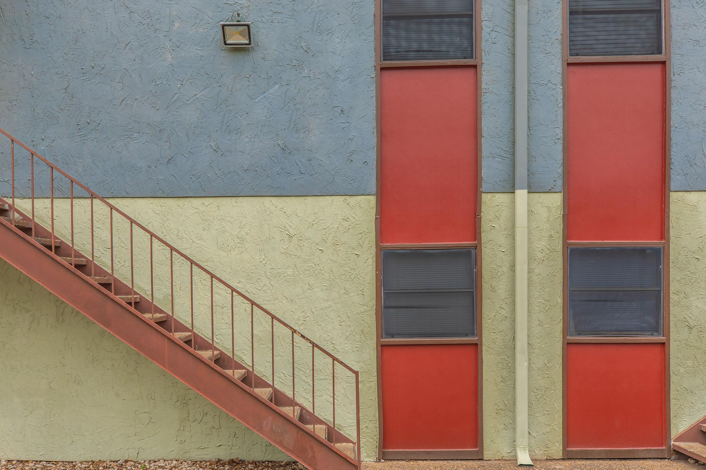 A modern building facade featuring a blue wall with green and beige sections, accented by two vertical red panels. A staircase with metal railings leads upward on the left, and a small light fixture is mounted above the doors. The overall aesthetic is clean and minimalistic.