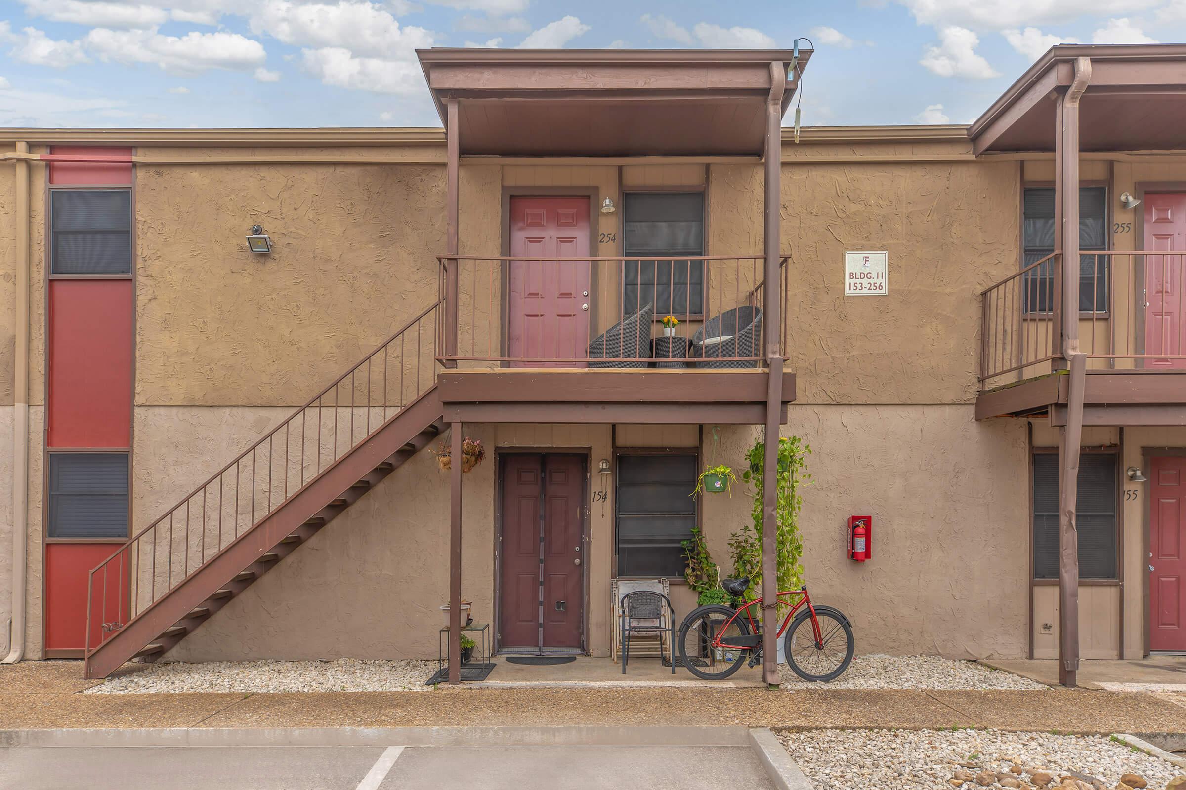 A two-story apartment building with a beige exterior. The lower level features a brown door with a small potted plant and a chair outside. A staircase leads to the upper level, which has a red door. A bicycle is parked nearby, and there’s a fire alarm box mounted on the wall. Several parking spaces are visible in front.