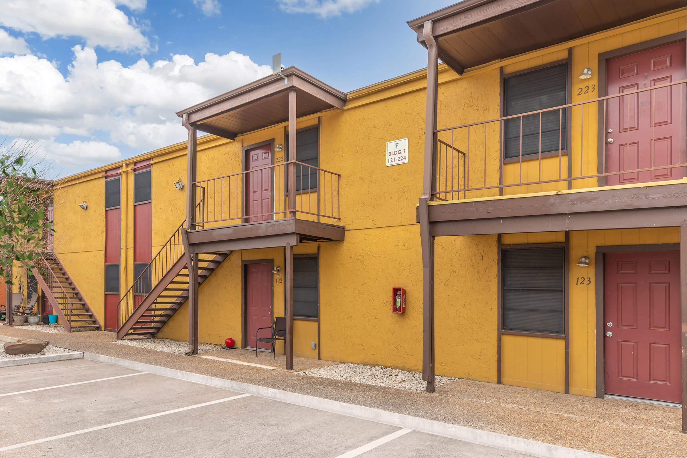 Two-story apartment building with a yellow exterior. There are two sets of red doors visible, labeled 123 and 223. Stairs lead to an upper level. The area is paved with a gravel pathway and a small sitting area visible. The sky is partly cloudy.