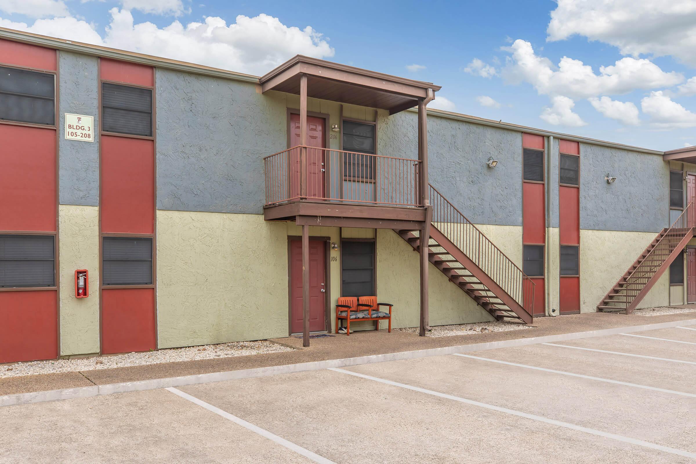 Exterior view of a two-story apartment building featuring a mix of red, green, and gray siding. It has a second-floor balcony with stairs leading up to it. In front, there is a small seating area with a bench, and several parking spaces visible in the foreground. The sky is partly cloudy.