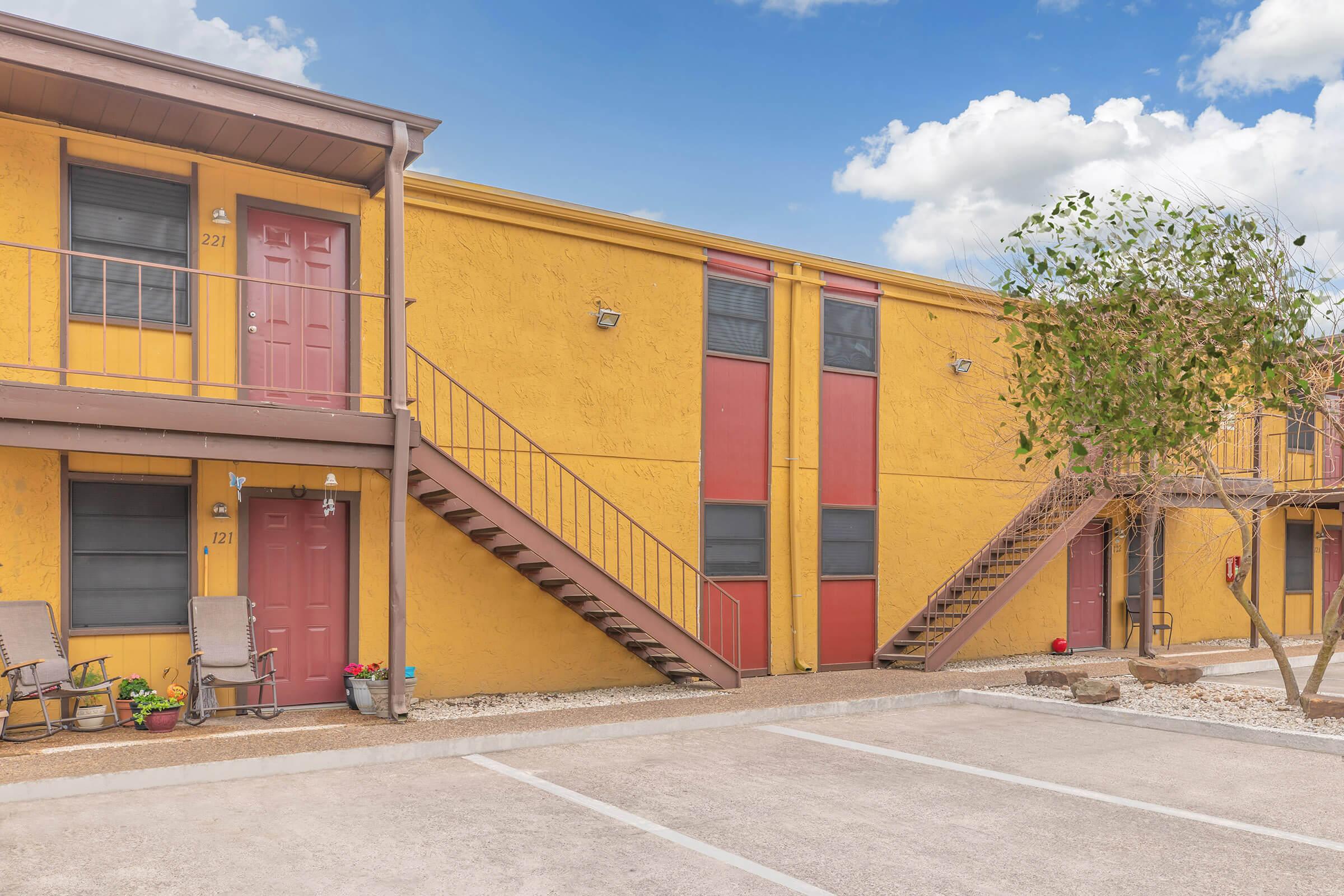 Two-story apartment building with yellow exterior and red doors. There are two sets of stairs leading to the upper level. Potted plants are placed in front of the ground-level units, and a small tree is nearby. The sky is blue with a few clouds, and there is a parking area in front.
