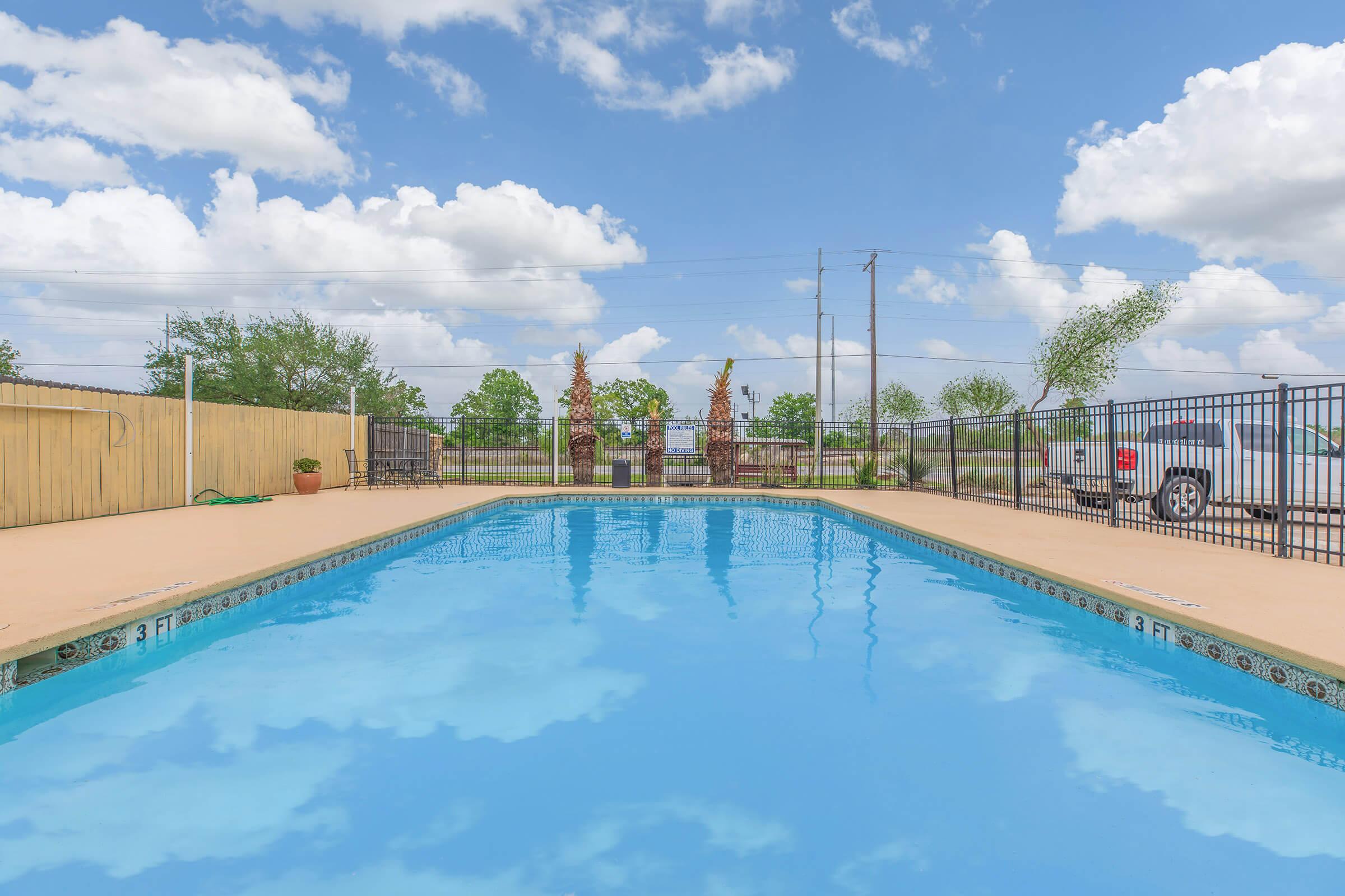 A clear blue swimming pool surrounded by a fence, with two decorative statues at one end. Lush greenery and clouds can be seen in the background, along with a vehicle parked nearby. The pool area is well-lit, suggesting a sunny day.
