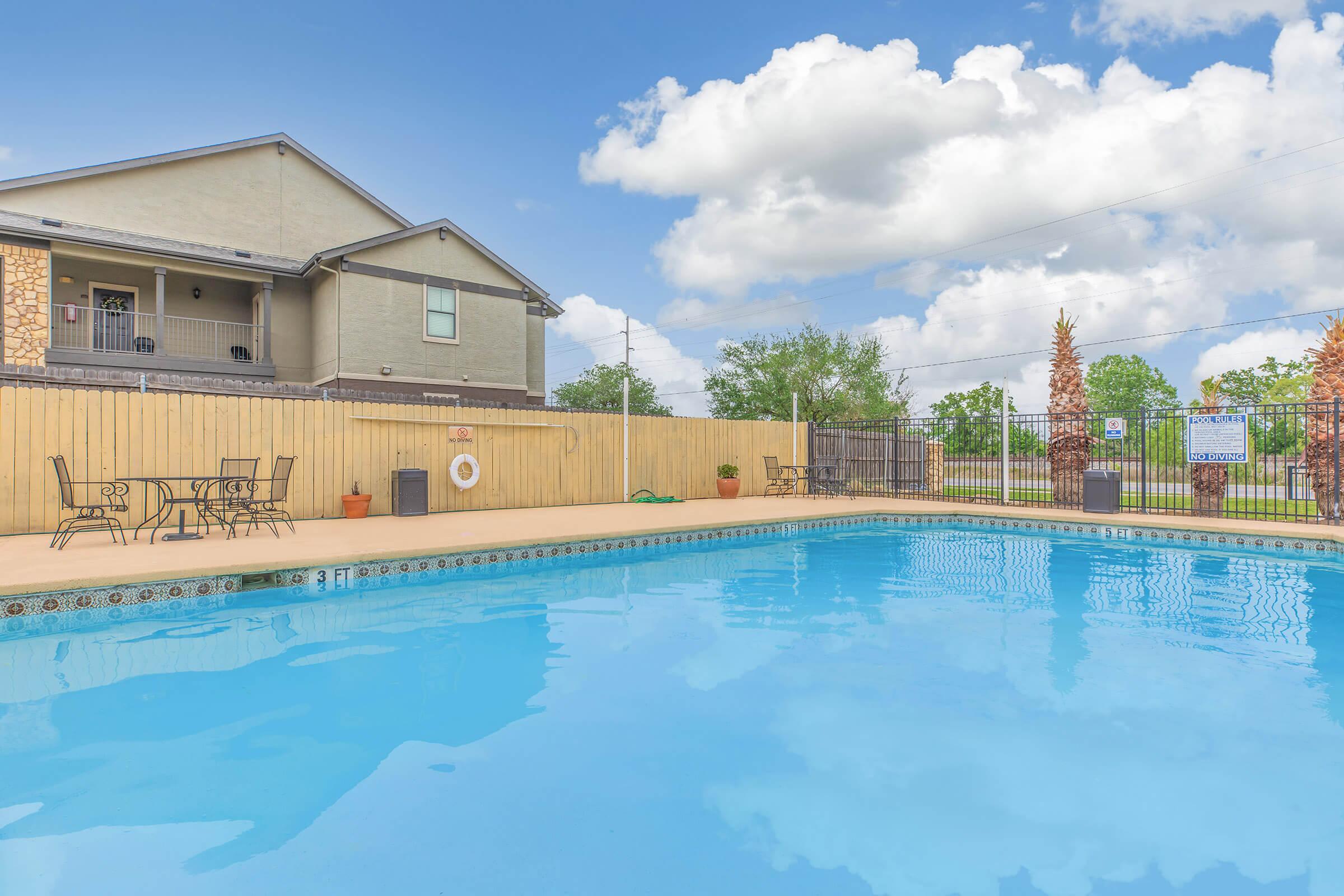 A clear blue swimming pool surrounded by a wooden fence, with lounge chairs and small tables nearby. In the background, there is a multi-story building and green trees under a partly cloudy sky.