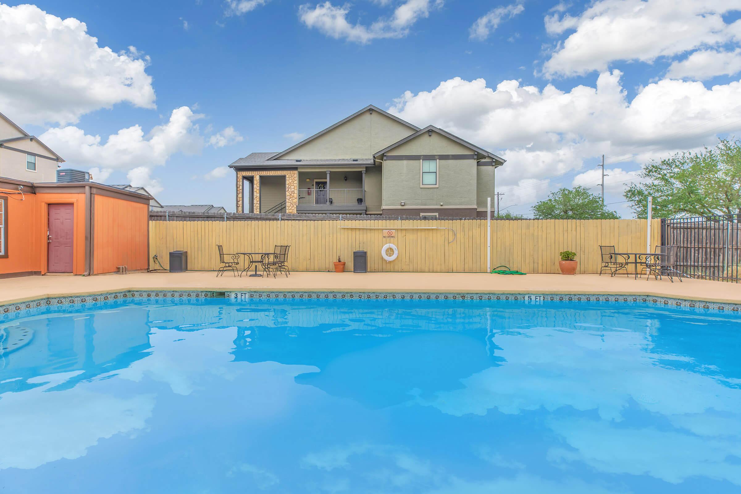 A clear swimming pool with blue water reflecting the sky and clouds. Surrounding the pool are patio areas with tables and chairs. In the background, a two-story house with a wooden fence is visible, along with trees and a partly cloudy sky.