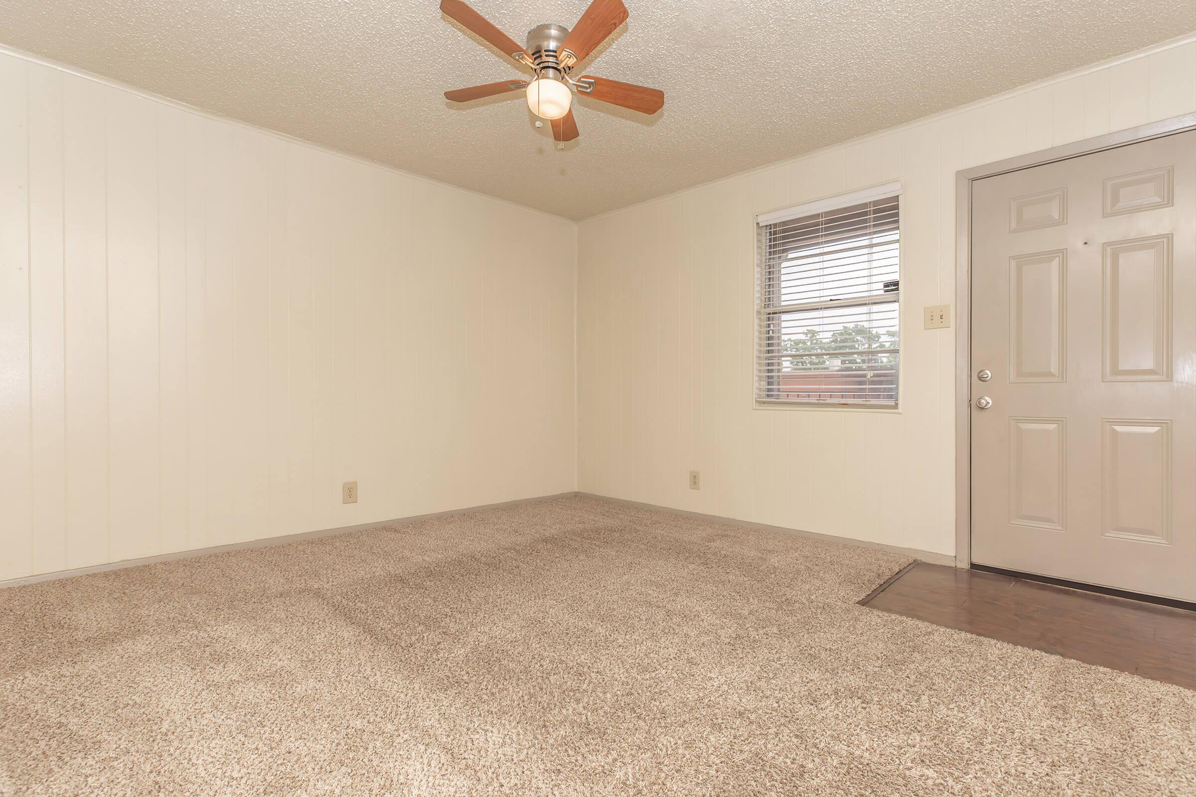 A spacious, empty room featuring beige walls, a ceiling fan with wooden blades, and cream-colored carpet. A window with blinds allows natural light, and a light brown door is visible on the right, leading to an exterior entry. The overall atmosphere is bright and inviting, perfect for furnishing.