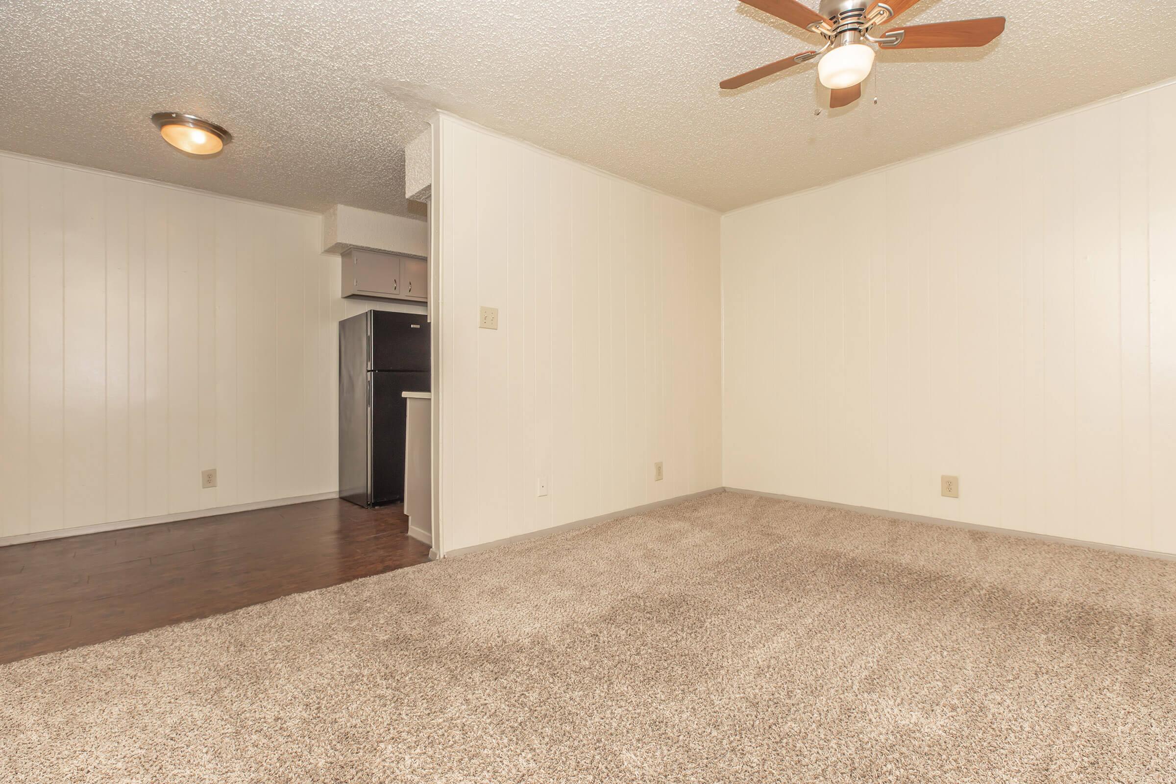 A spacious, empty living room with beige walls and carpet. There is a ceiling fan, and a glimpse of a kitchen area with dark appliances visible in the background. The room is well-lit, creating a warm and inviting atmosphere.