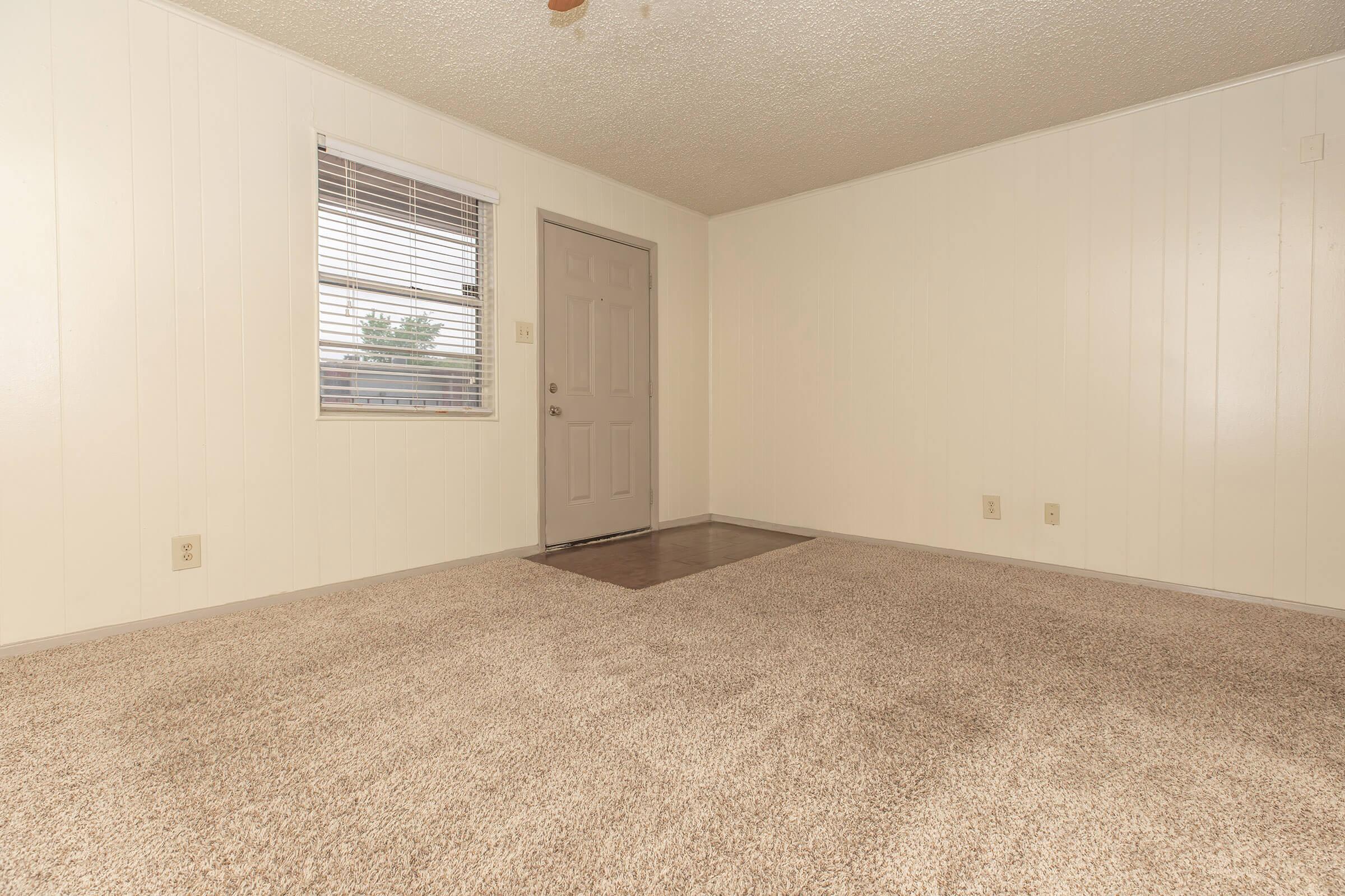 Empty room with beige carpet, light-colored walls, and a window with horizontal blinds. A closed door is visible on one wall, with a small beige mat near it. The space is well-lit and has a ceiling fan. The overall ambiance is simple and clean, suitable for personalization.