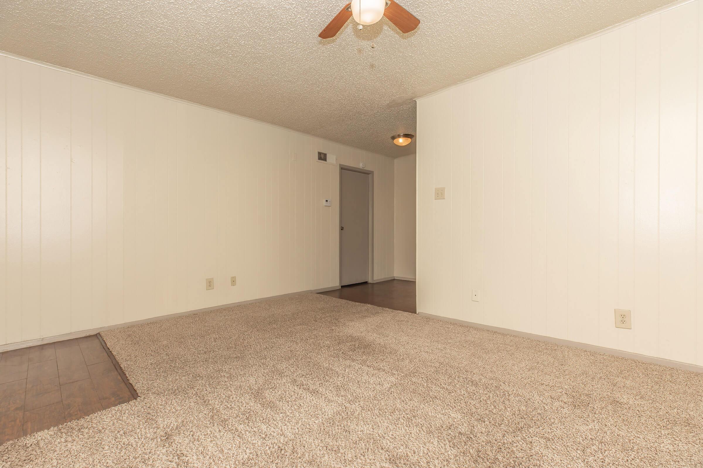 A spacious, empty room with light-colored walls and a ceiling fan. The floor features a section of carpet, with a smooth, dark wood floor visible at the edge. A doorway leads to another room, and there is a light fixture on the ceiling, providing soft illumination.