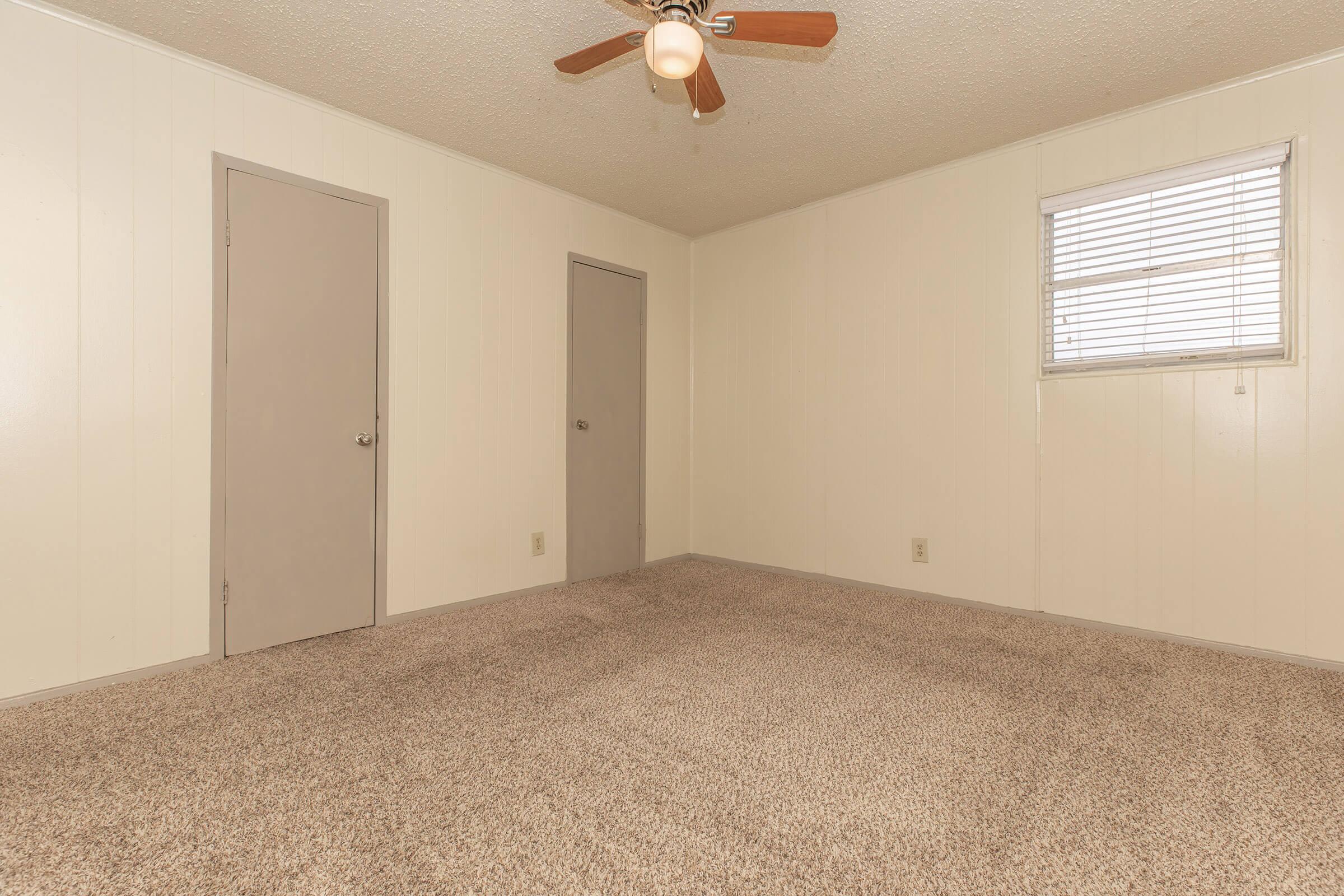 Empty room with light beige walls, a ceiling fan, and carpeted floor. Two gray doors are visible, leading to closets or other spaces. A small window with blinds allows natural light to enter. The overall appearance is clean and simple, suitable for various uses or potential decoration.