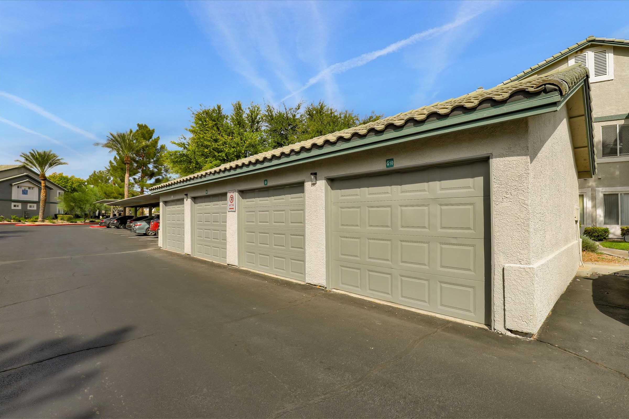 A row of closed garage doors in a residential area, with a paved driveway. Lush greenery and palm trees are visible in the background, alongside parked cars. The sky features light clouds and a bright blue color, indicating a clear day.