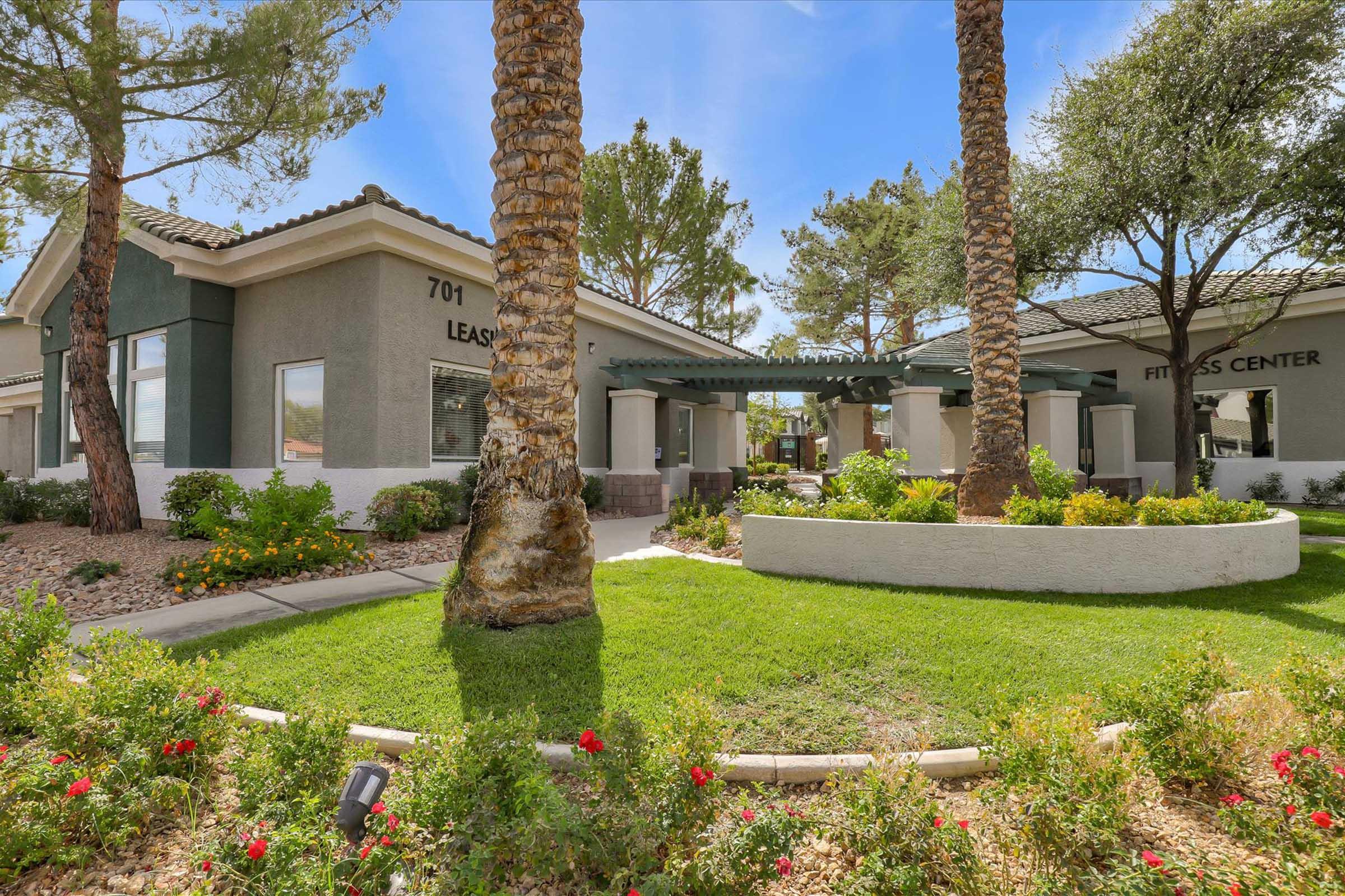 A modern building with a leasing office and fitness center, surrounded by lush greenery and colorful flower beds. Palm trees and other plants add to the inviting landscape. The sky is clear and blue, enhancing the overall appealing atmosphere of the property.
