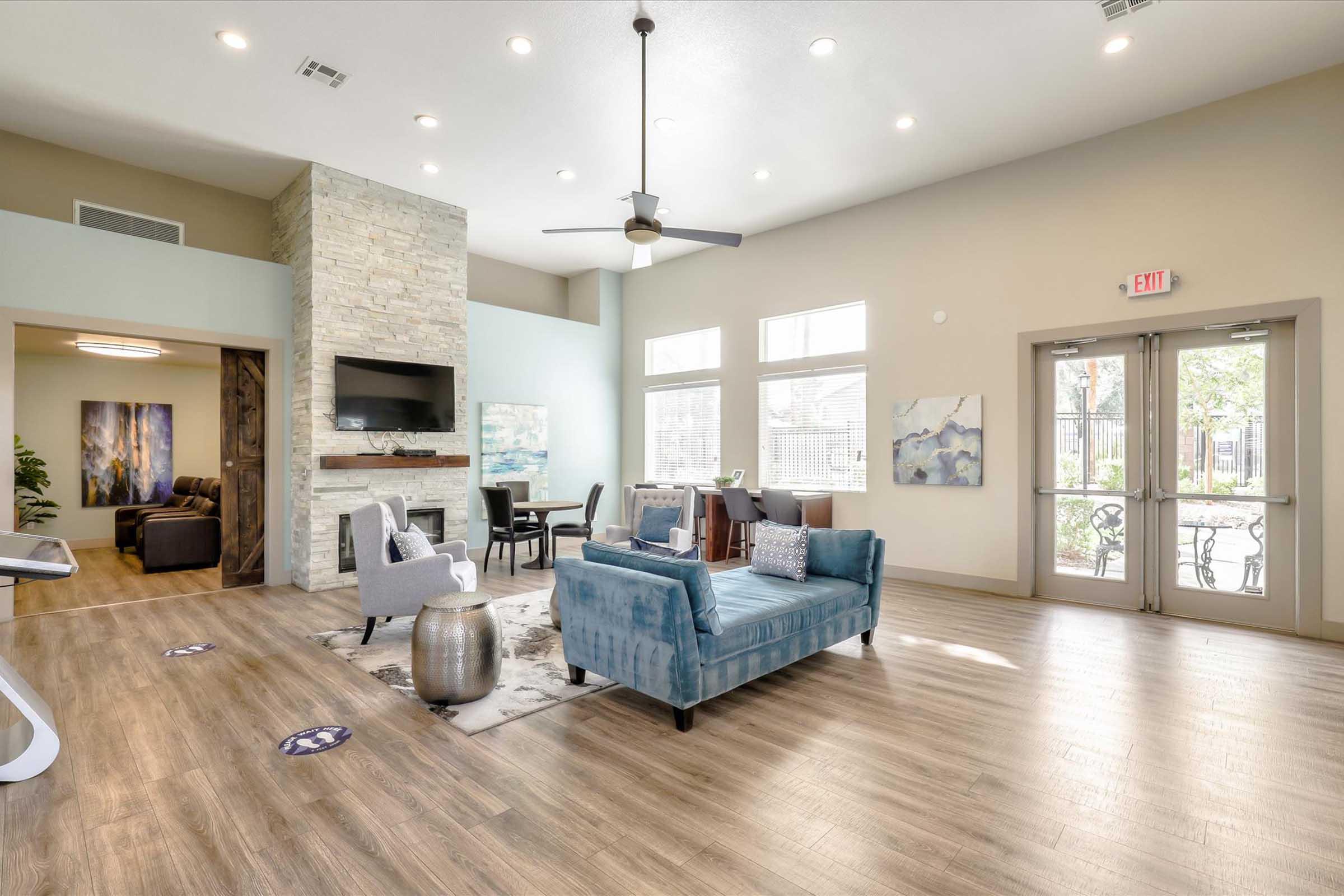 A spacious and modern lounge area featuring a blue sofa, a coffee table, and a stone fireplace. Large windows allow natural light, and there are chairs and a dining table. Decor includes artwork on the walls, and the flooring is wooden. An exit door leads to an outdoor area.