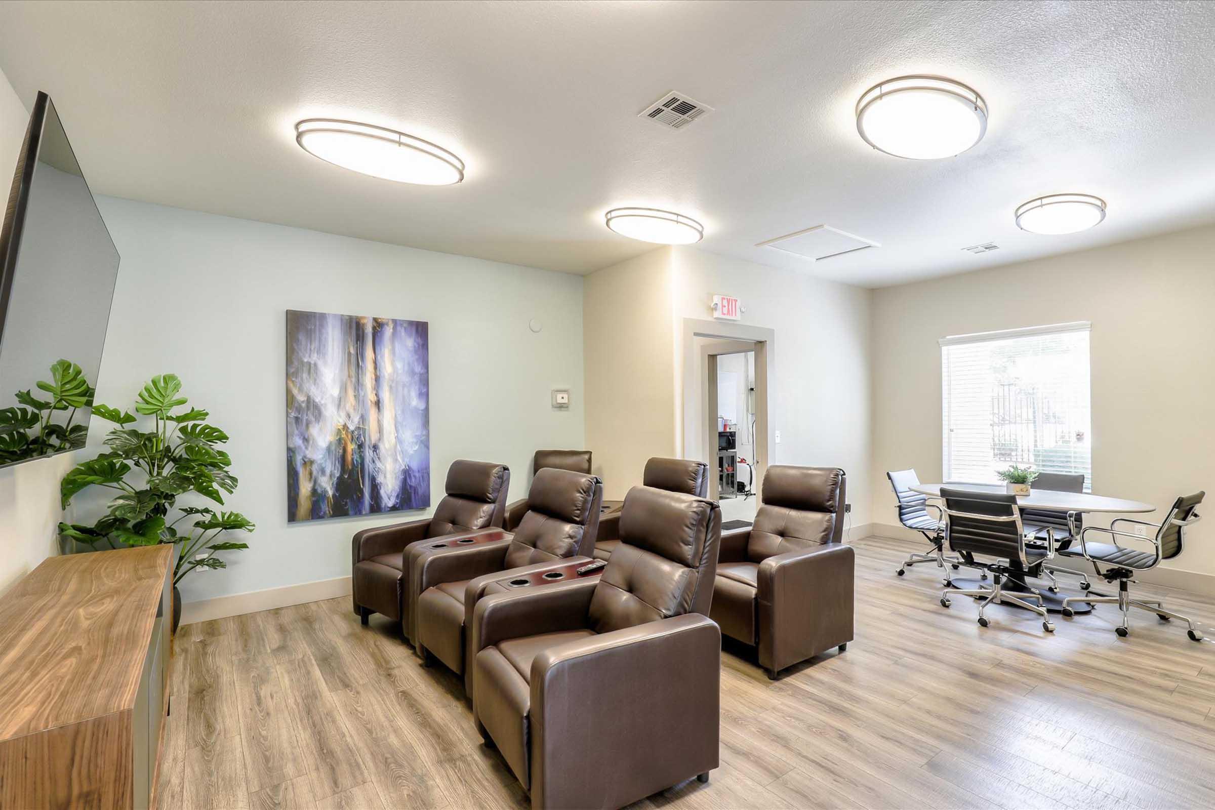 A modern waiting or lounge area featuring comfortable leather recliners with cup holders, a large wall-mounted TV, a decorative wall painting, and a small meeting area with a table and chairs. Natural light flows in through a window, complemented by potted plants.
