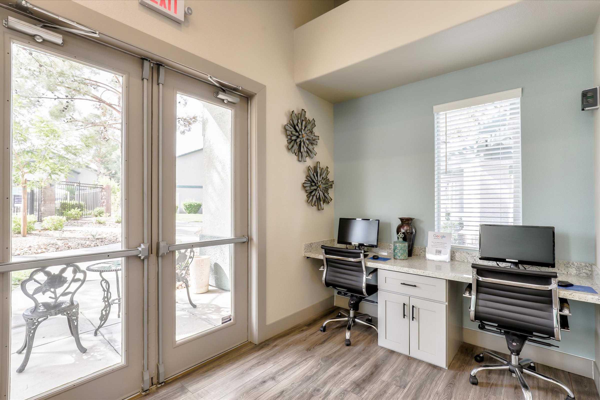 A well-lit reception area featuring two black office chairs at desks with monitors. The space includes large glass doors leading outside, decorative wall pieces, and large windows with natural light illuminating the room. Lush greenery is visible through the doors, enhancing the welcoming atmosphere.