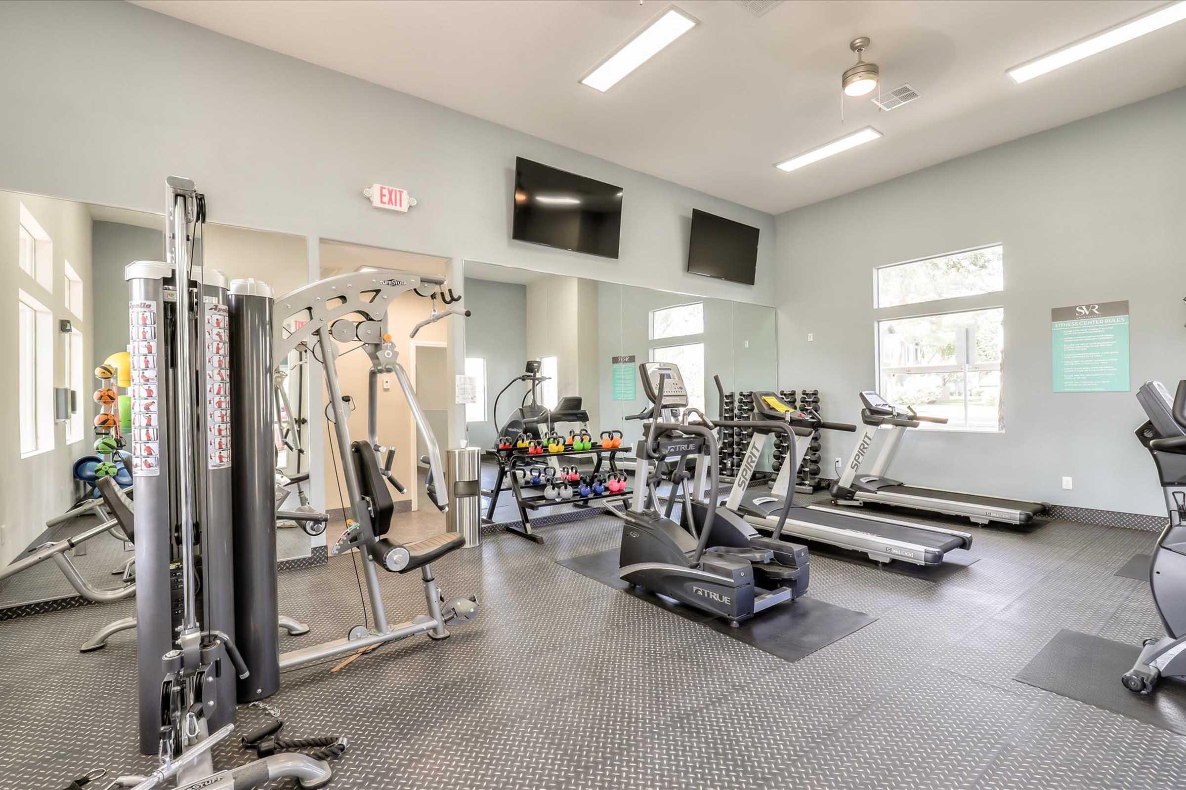 Interior view of a modern gym featuring various exercise equipment including weight machines, treadmills, and free weights. The bright, spacious room has large windows and wall-mounted TVs, creating a motivating workout environment. Rubber flooring enhances safety and comfort during workouts.