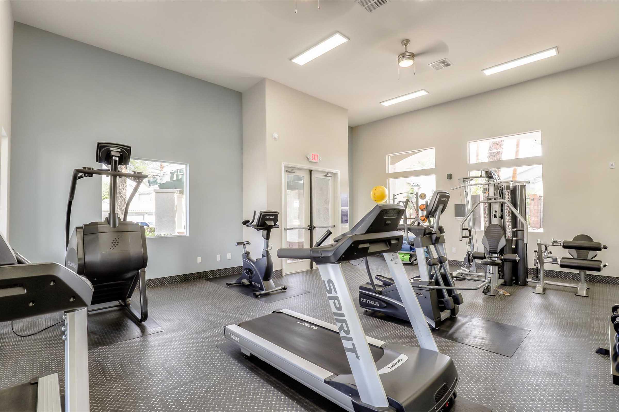 A well-lit gym with various workout equipment including treadmills, stationary bikes, and strength training machines. The space features large windows allowing natural light, and the flooring is black rubber mats.