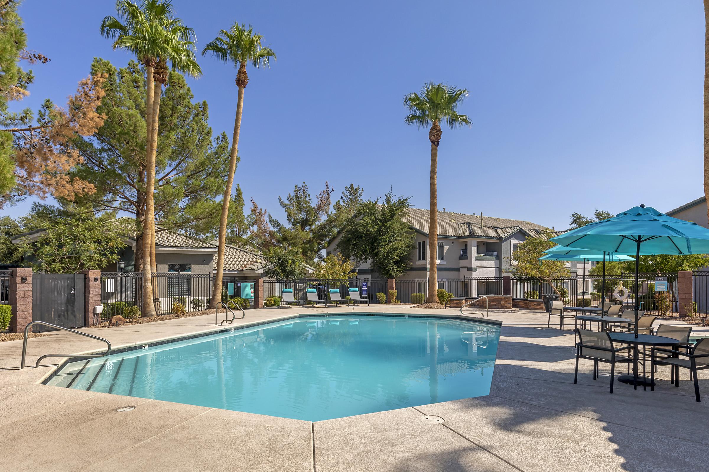 A sunny pool area featuring a clear blue swimming pool surrounded by palm trees. There are lounge chairs and an umbrella for shade, along with tables and chairs for seating. In the background, apartment buildings and well-maintained landscaping complete the inviting atmosphere.