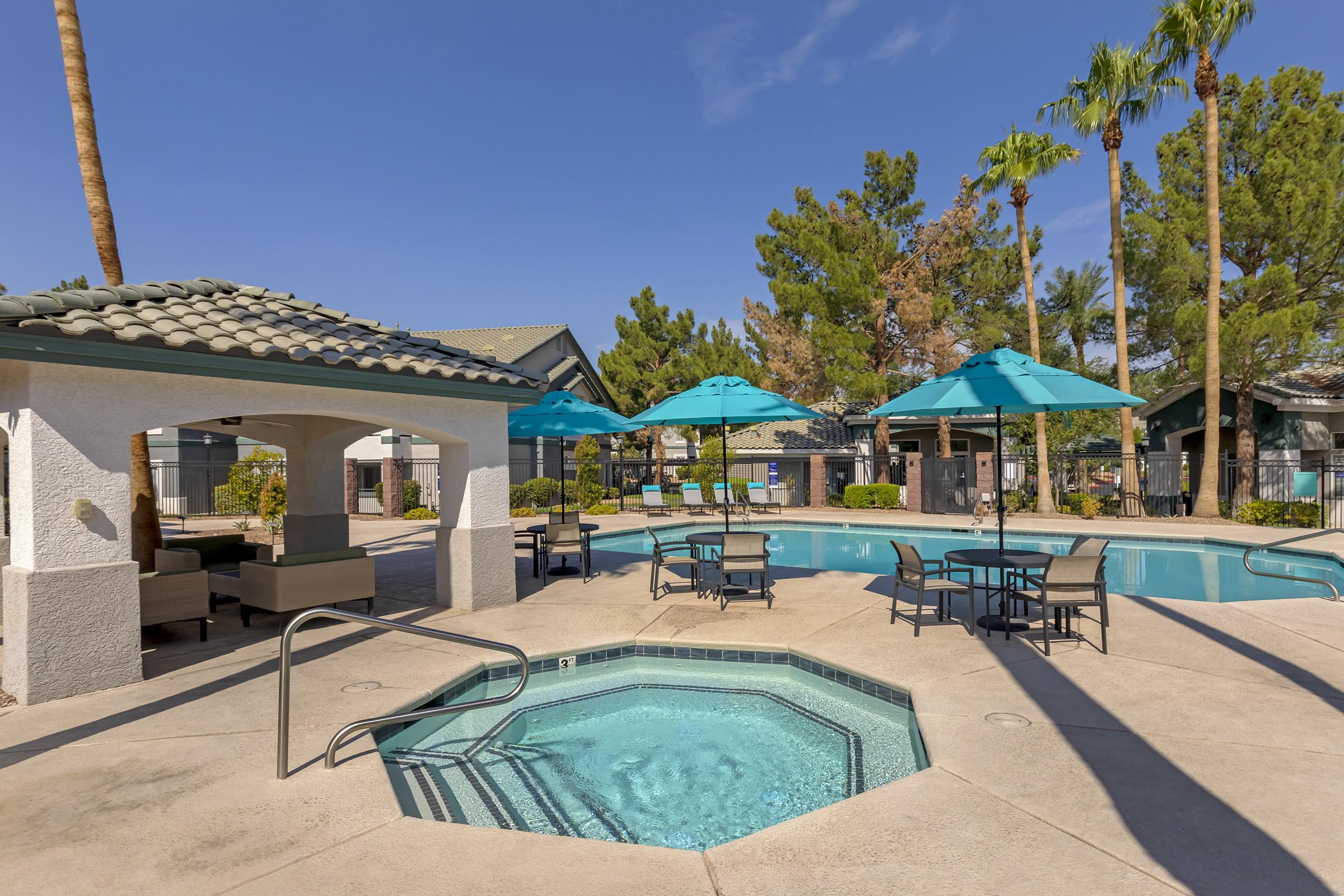 A serene outdoor pool area featuring a hot tub, lounge chairs, and turquoise umbrellas. Palm trees surround the space, and nearby buildings are partially visible under a clear blue sky. The setting invites relaxation and enjoyment, perfect for leisure activities.