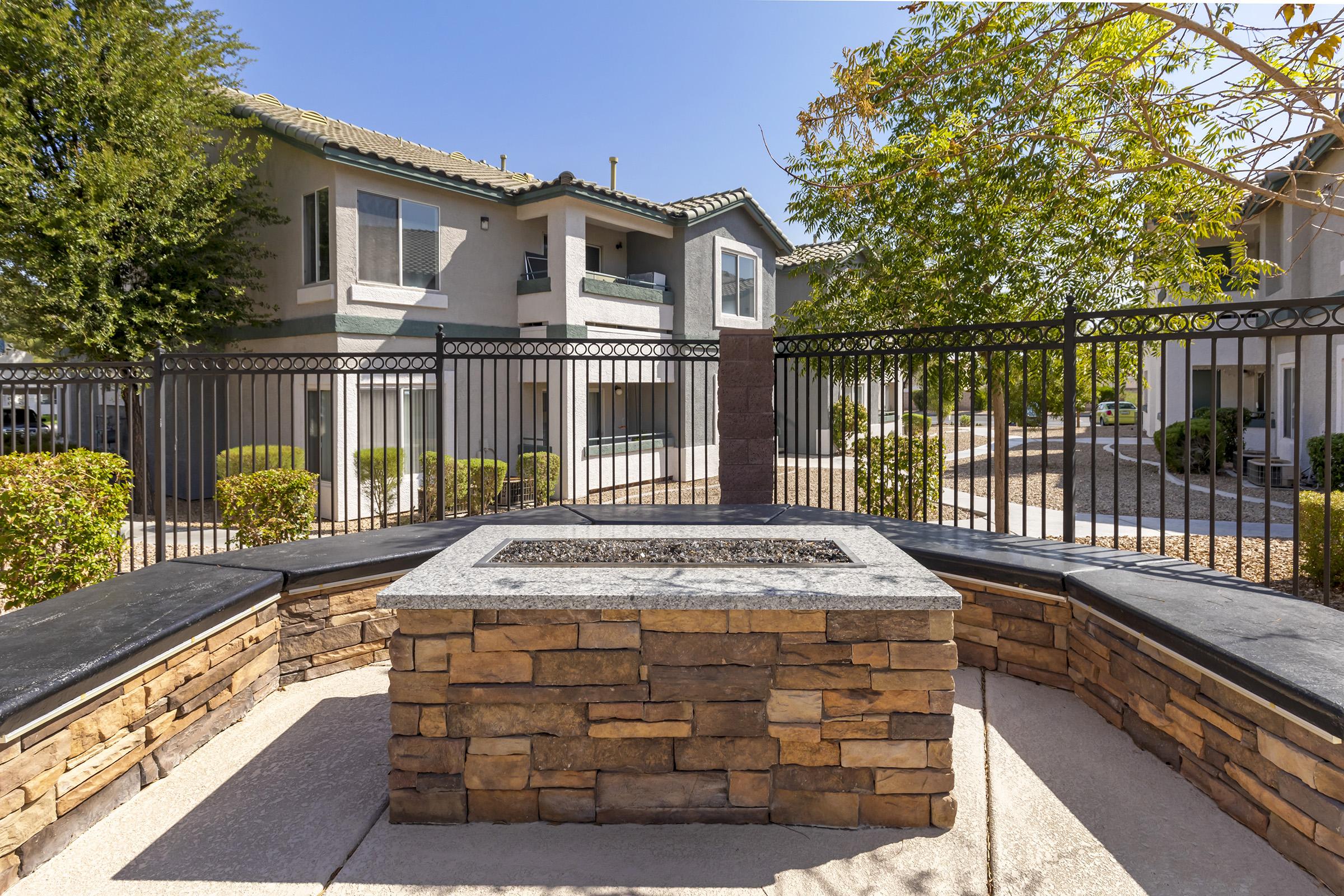 Stone fire pit surrounded by a circular seating area, with a background of two residential buildings and a green tree. The scene is bright and sunny, showcasing a landscaped outdoor area, enclosed by a decorative black metal fence.