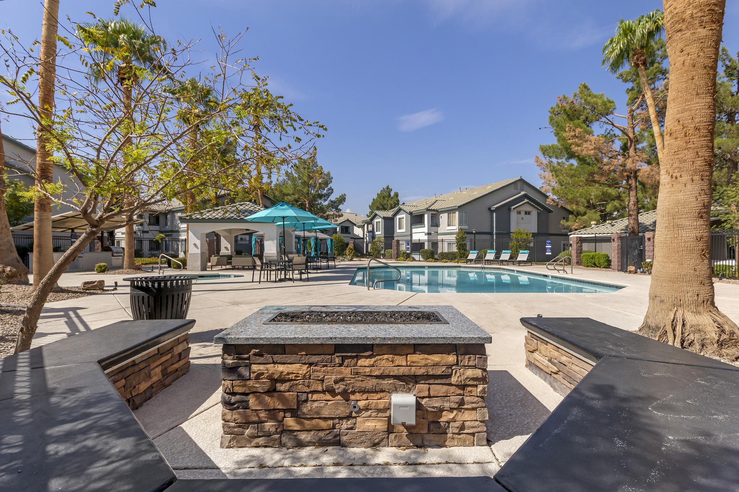 A serene outdoor area featuring a swimming pool surrounded by lounge chairs, palm trees, and a gazebo. A stone fire pit with seating is in the foreground, creating a relaxing atmosphere. Bright blue skies reflect a sunny day, enhancing the inviting ambiance of this recreational space.