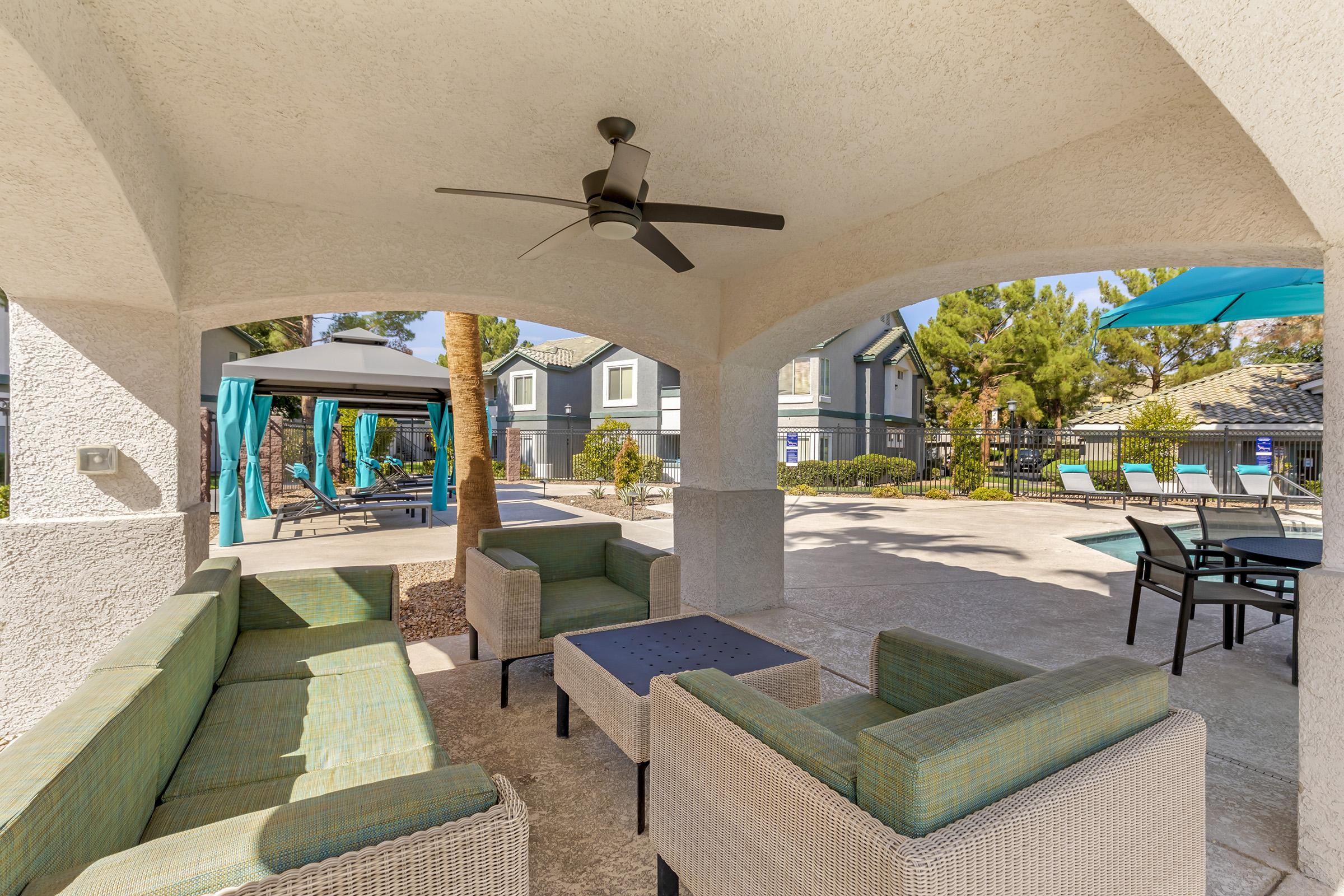 A shaded outdoor lounge area featuring modern wicker furniture with blue and green cushions. A ceiling fan hangs above, and in the background, there are palm trees, a pool area, and blue umbrellas beside lounge chairs, creating a relaxing atmosphere.