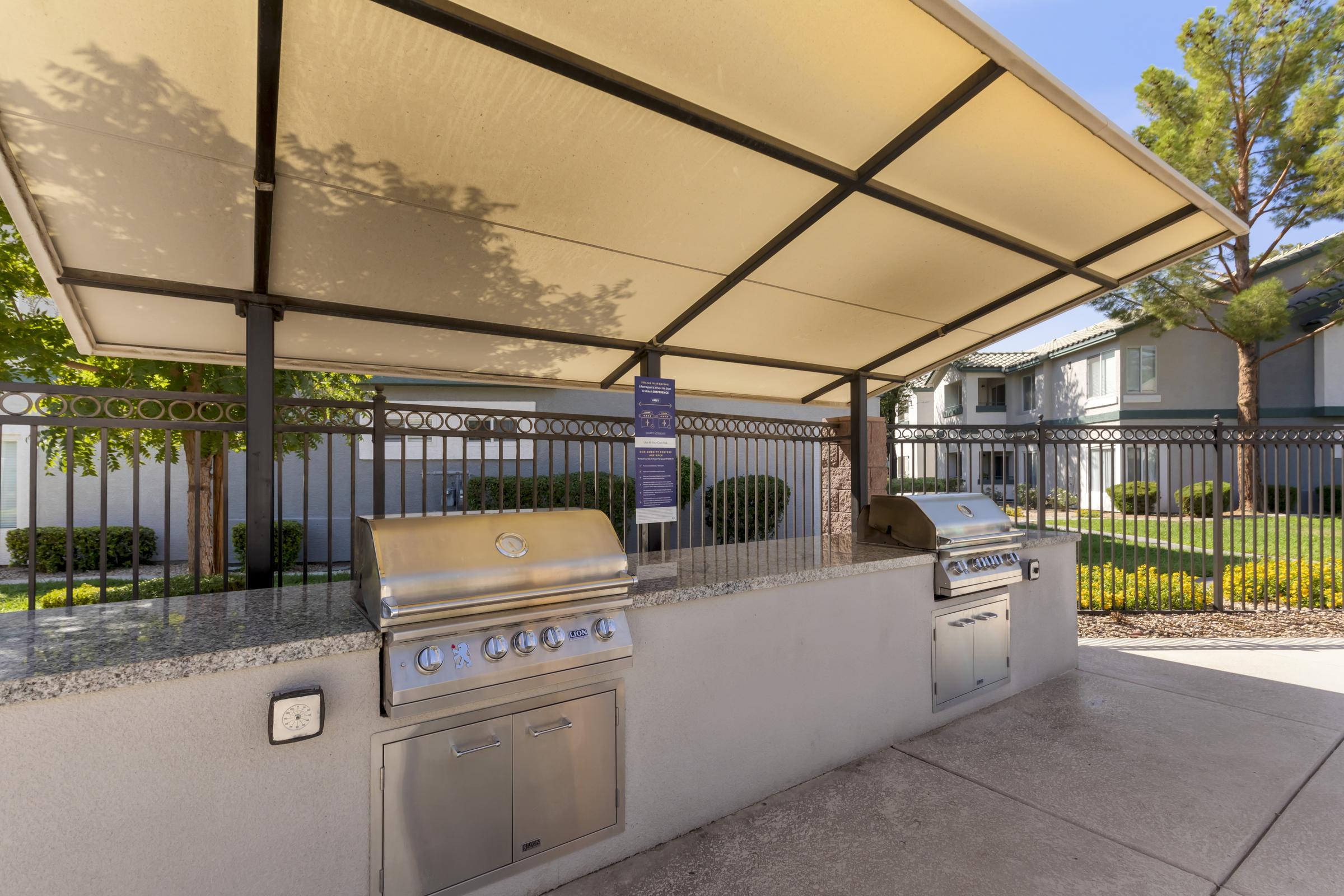 Outdoor grilling area featuring two stainless steel barbeque grills under a shaded pavilion. The space is surrounded by green landscaping and a fence, with a building visible in the background. Ideal for social gatherings and cooking outdoors.