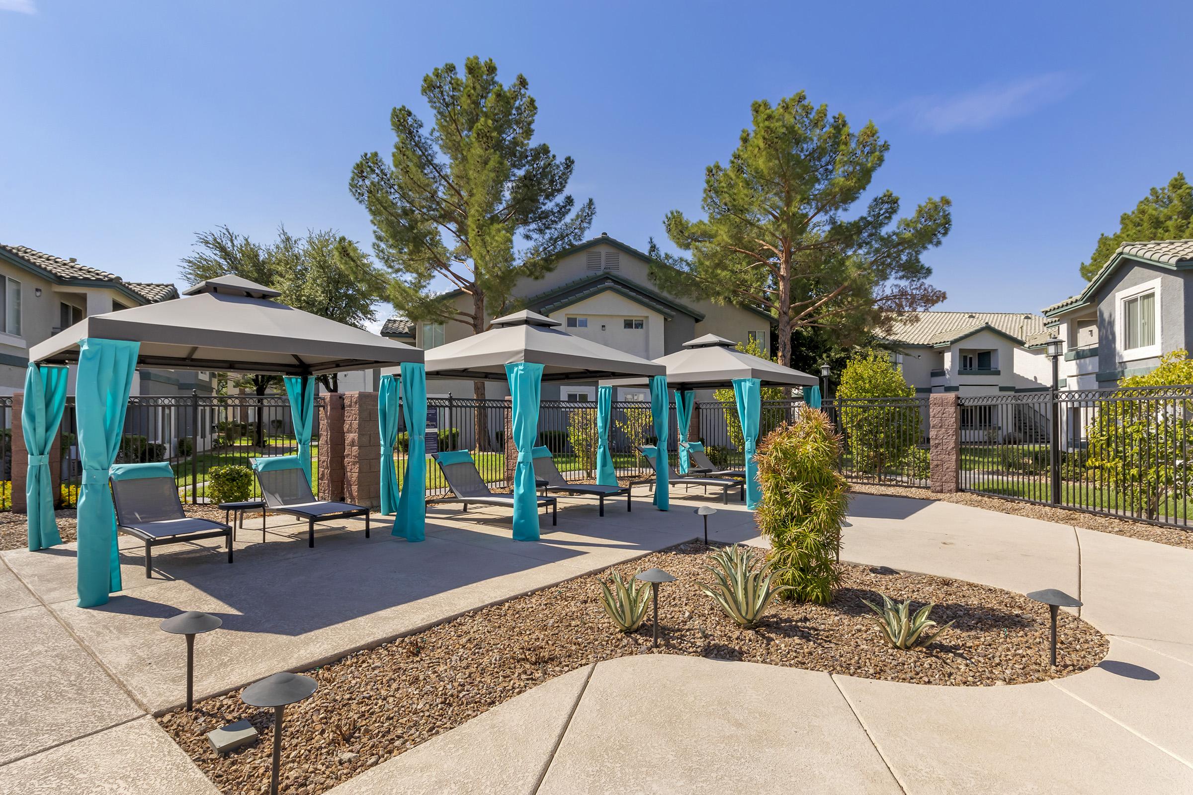 A sunny outdoor area featuring several shaded cabanas with turquoise curtains, lounge chairs, and landscaped gravel. Surrounding greenery includes trees and shrubs, while a fence encloses the space, providing a relaxing atmosphere for leisure and enjoyment.