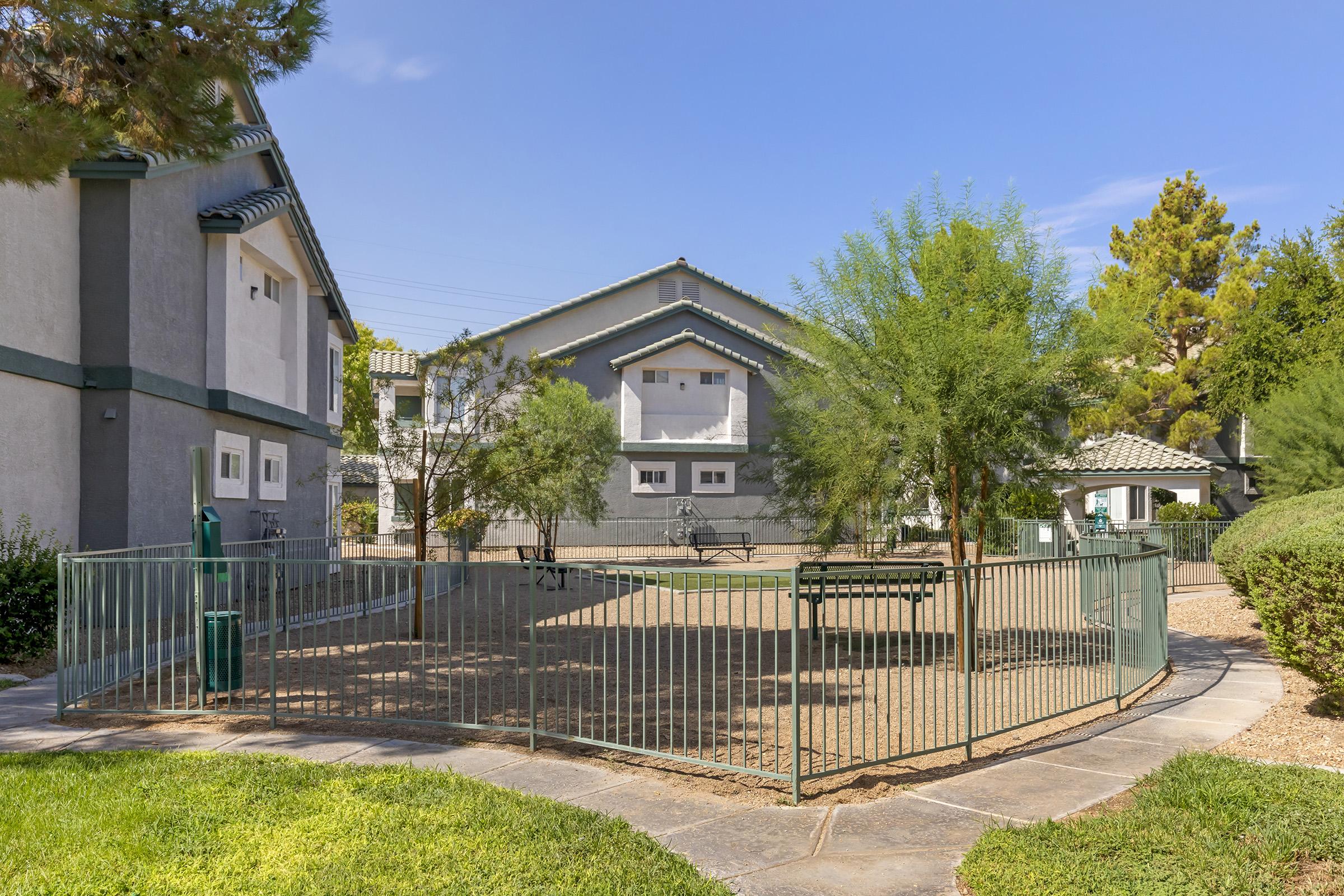 A residential area featuring a small playground enclosed by a fence. Nearby, there are two apartment buildings with greenery and trees. The sky is clear and blue, creating a pleasant outdoor atmosphere.