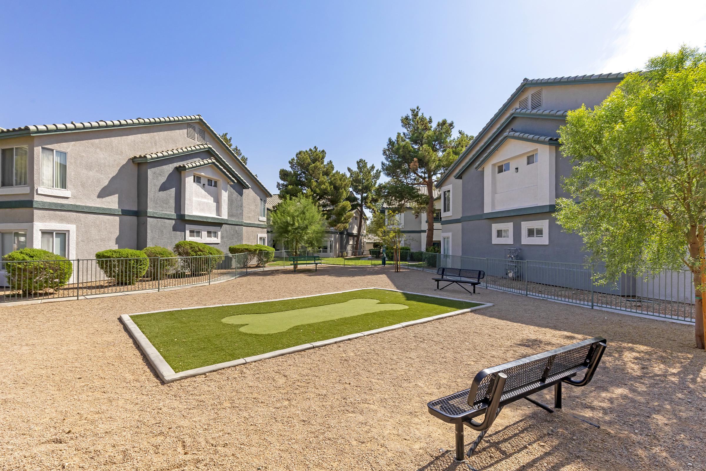 A quiet courtyard surrounded by two apartment buildings. There is a green, artificial turf area shaped like a footprint in the center, bordered by gravel. A black bench sits along one side, and there are small shrubs and trees throughout the space, providing a serene environment for relaxation.