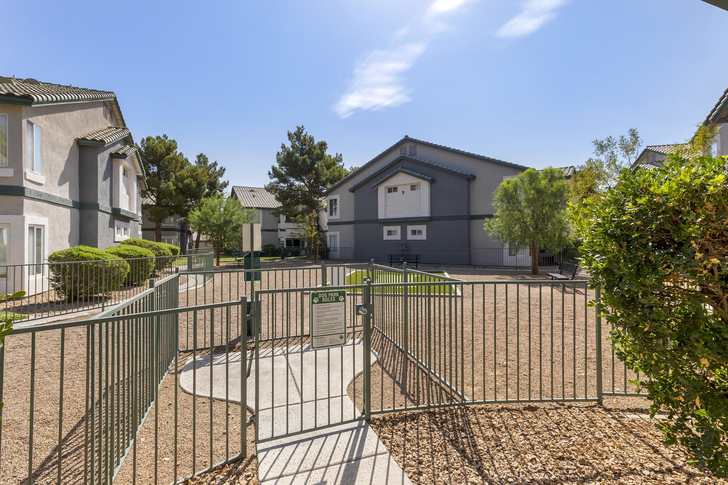 A view of a residential area featuring two-story buildings with green landscaping. A fenced area with a pathway leads into a courtyard with gravel and shrubs. Clear blue skies are visible above, suggesting a sunny day.
