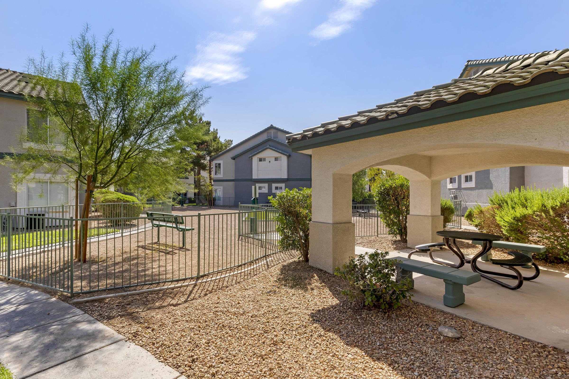 A fenced outdoor area featuring a picnic table and benches under a shaded gazebo. Surrounding the space are well-maintained shrubs and trees, with a glimpse of nearby buildings in the background. The sky is clear and sunny, creating a pleasant atmosphere for relaxation.