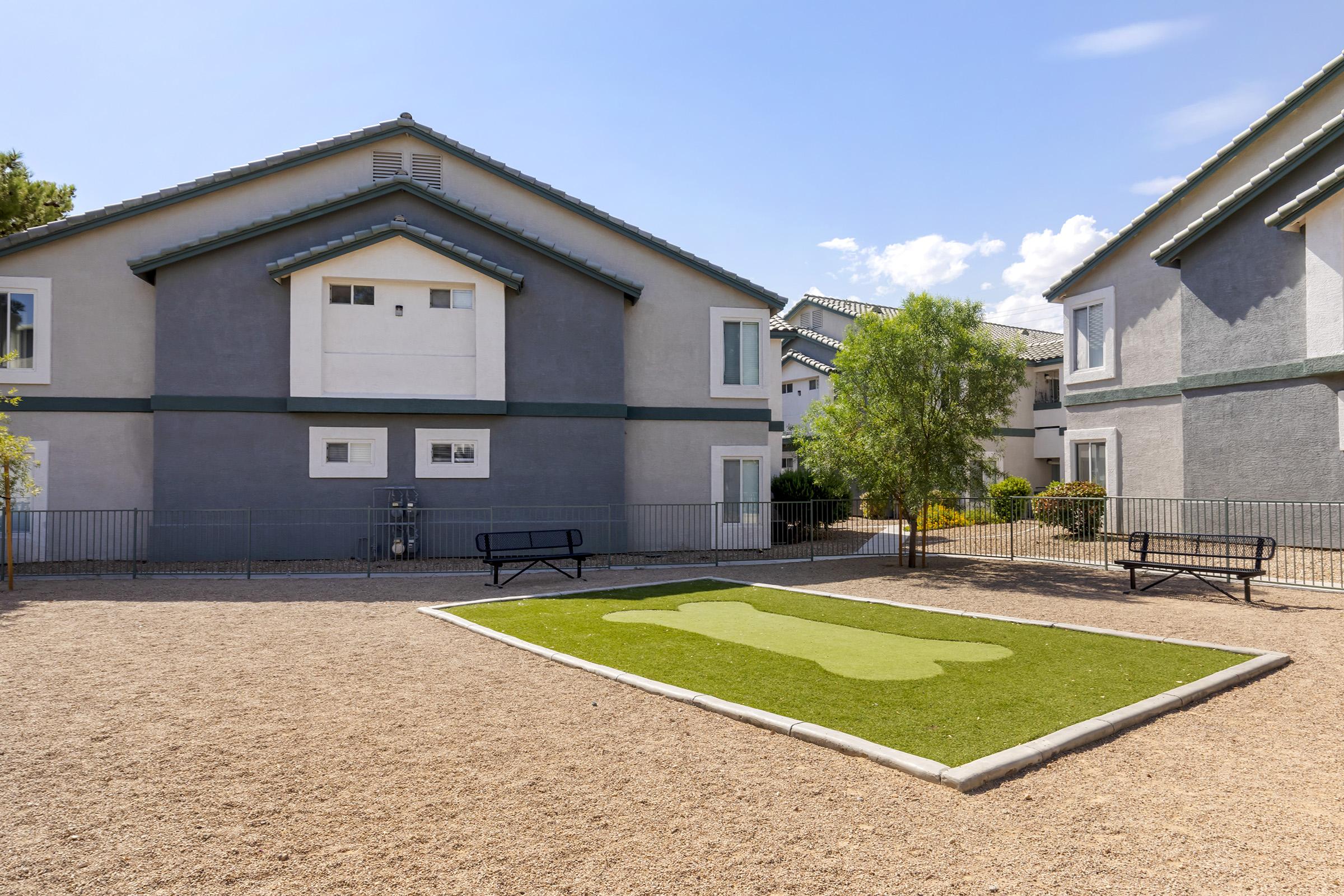 A view of an apartment complex featuring two buildings with a gravel courtyard. In the foreground is a green, artificial turf area shaped like a dog, bordered by a concrete edge. There are benches nearby, and trees and shrubs are visible in the surrounding landscape under a blue sky with a few clouds.