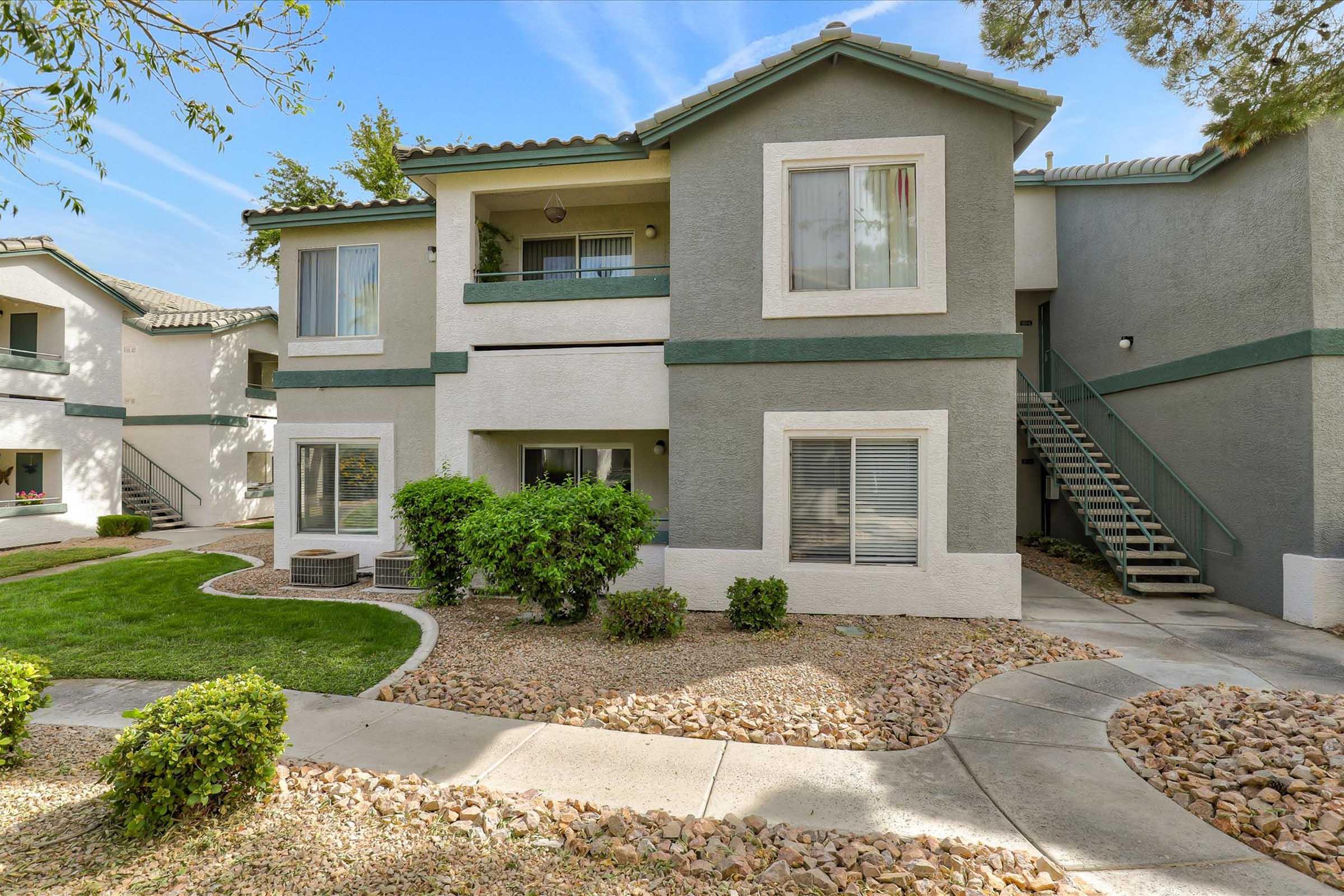 Two-story apartment building with a landscaped front yard featuring grass, bushes, and a stone pathway. The exterior has light gray and white walls, with windows on each level. A staircase leads to the second floor, and the surroundings include a well-maintained lawn and small rocks. Bright blue sky in the background.