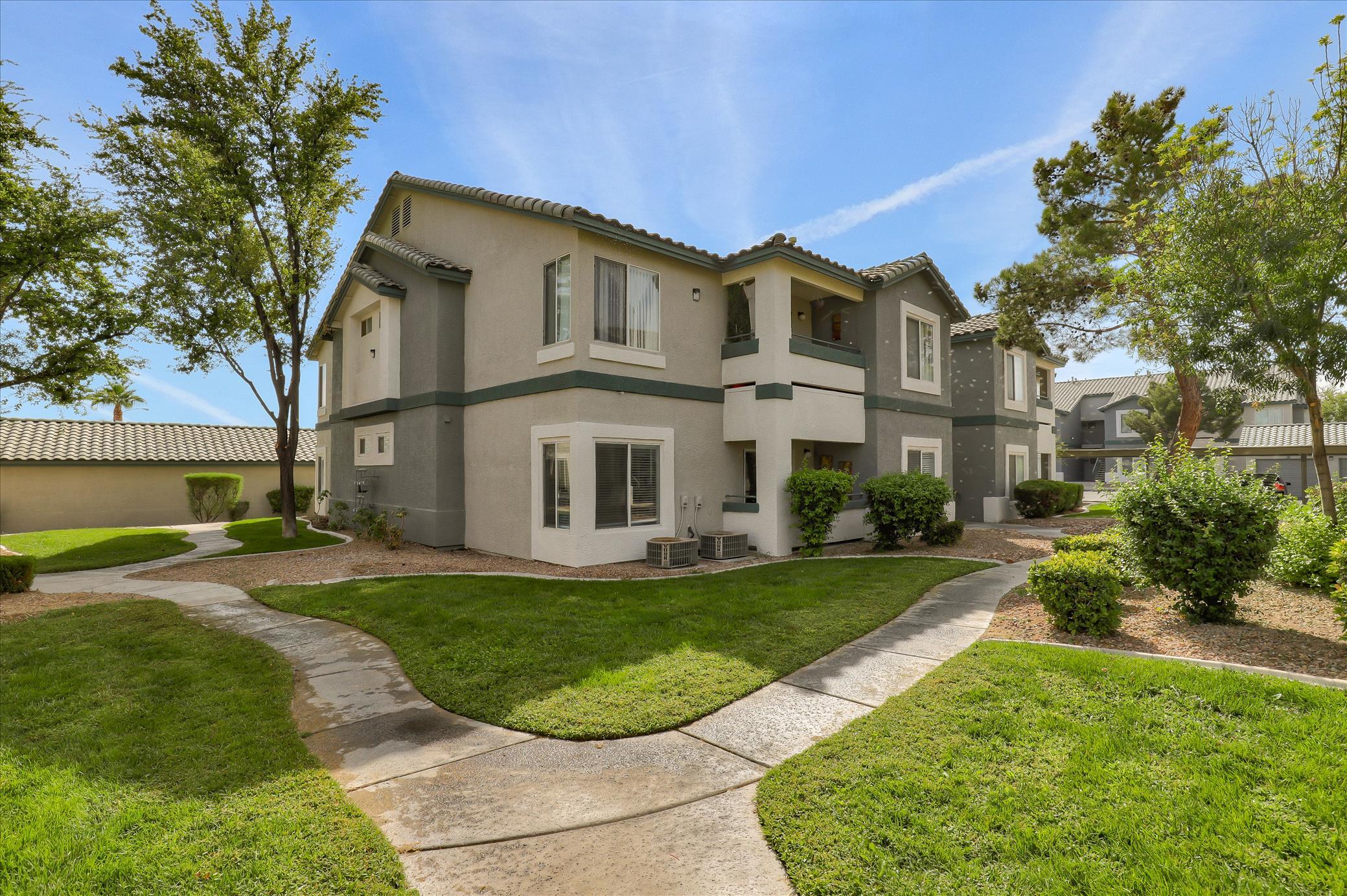 A landscaped apartment complex featuring two-story buildings with balconies, surrounded by green lawns and pathway walkways. Trees and shrubs enhance the outdoor space, creating an inviting residential environment. Bright blue sky with a few clouds above.