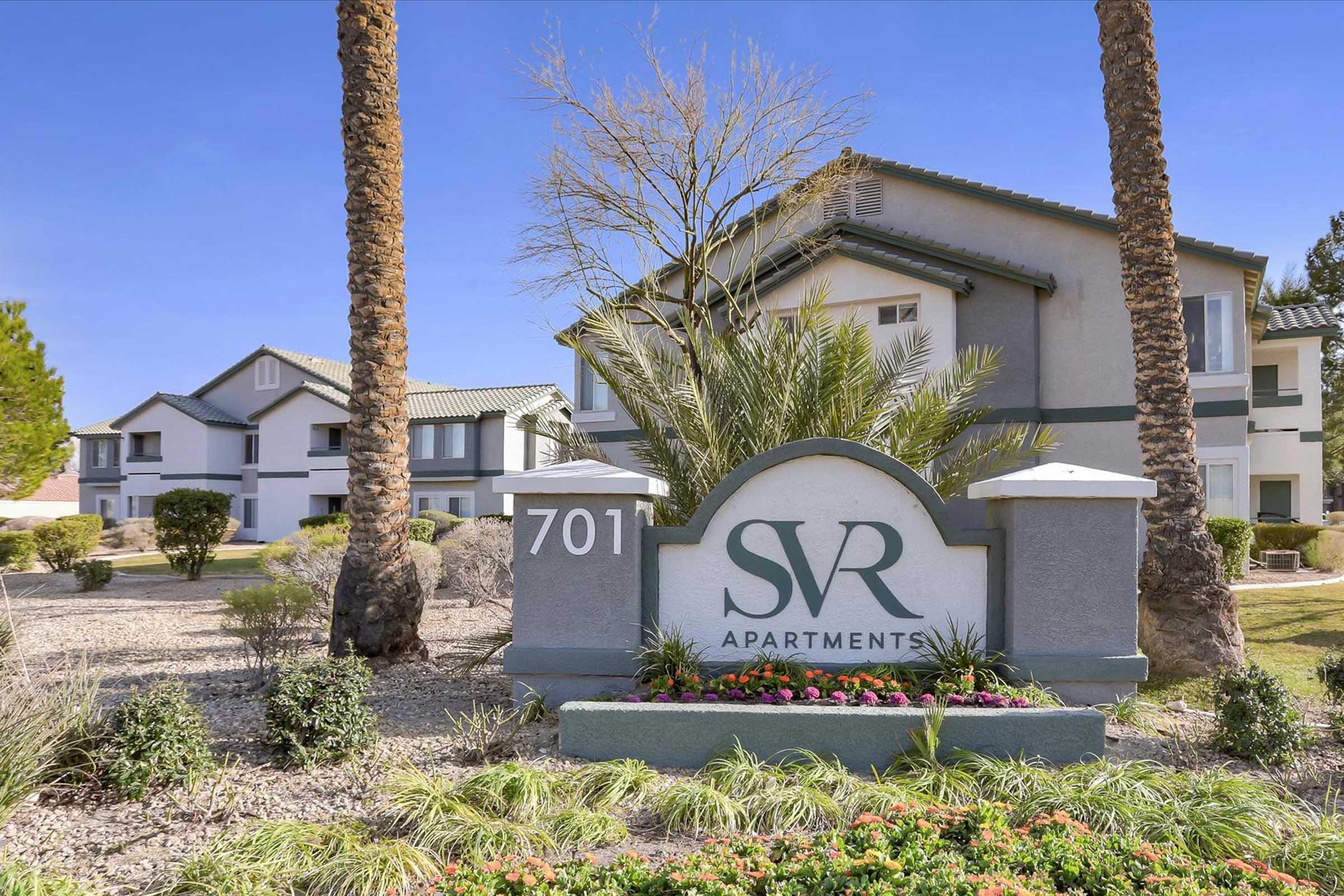 Sign for SVR Apartments with the number 701, surrounded by landscaped areas featuring palm trees and colorful flowers. In the background, two multi-story apartment buildings are visible against a blue sky.