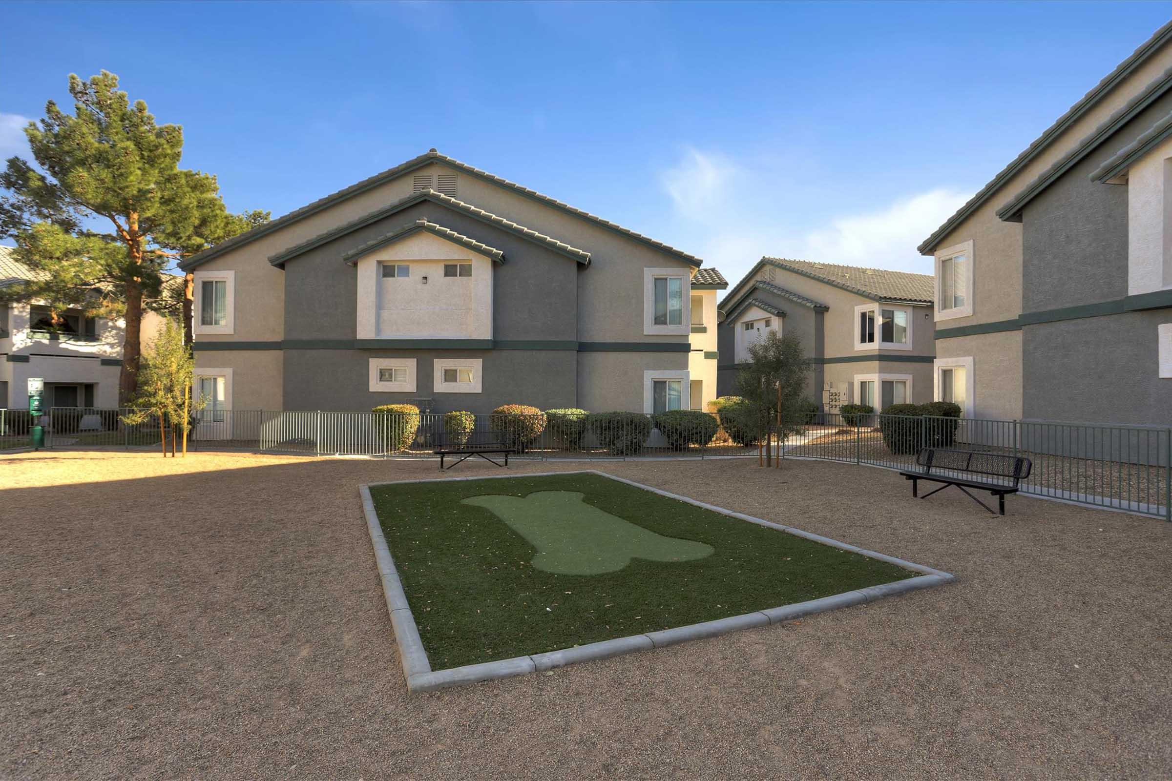 A landscaped courtyard featuring multiple two-story apartment buildings. The ground is covered in gravel, with a green area shaped like a large bone, likely intended for dogs. Shrubs are planted along the pathways, and a bench is visible nearby. Clear blue skies and a sunny atmosphere enhance the scene.