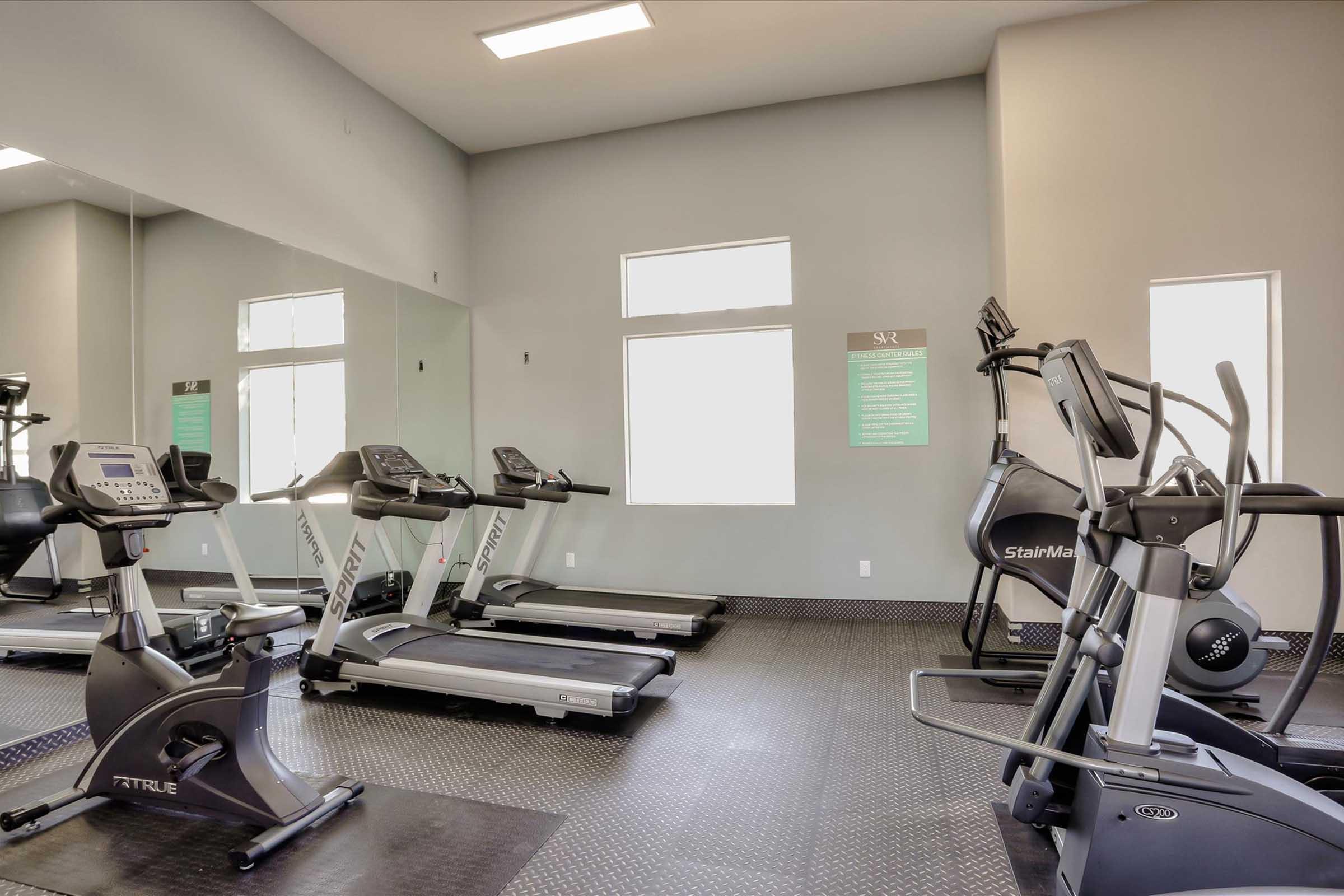 A modern gym interior featuring several exercise machines, including treadmills and an elliptical. The walls are painted in a neutral color, and there are large windows allowing natural light. Informational signs are displayed on the walls, and the flooring is dark, textured rubber.
