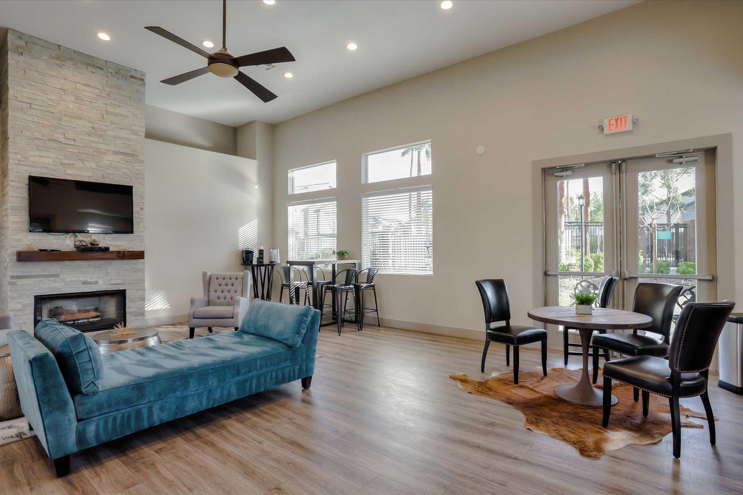 Modern living room with a light color scheme featuring a stone fireplace, a blue sofa, two gray armchairs, a round dining table with black chairs, and large windows allowing natural light. The space has wooden flooring and a cozy atmosphere, suitable for relaxation and gatherings.