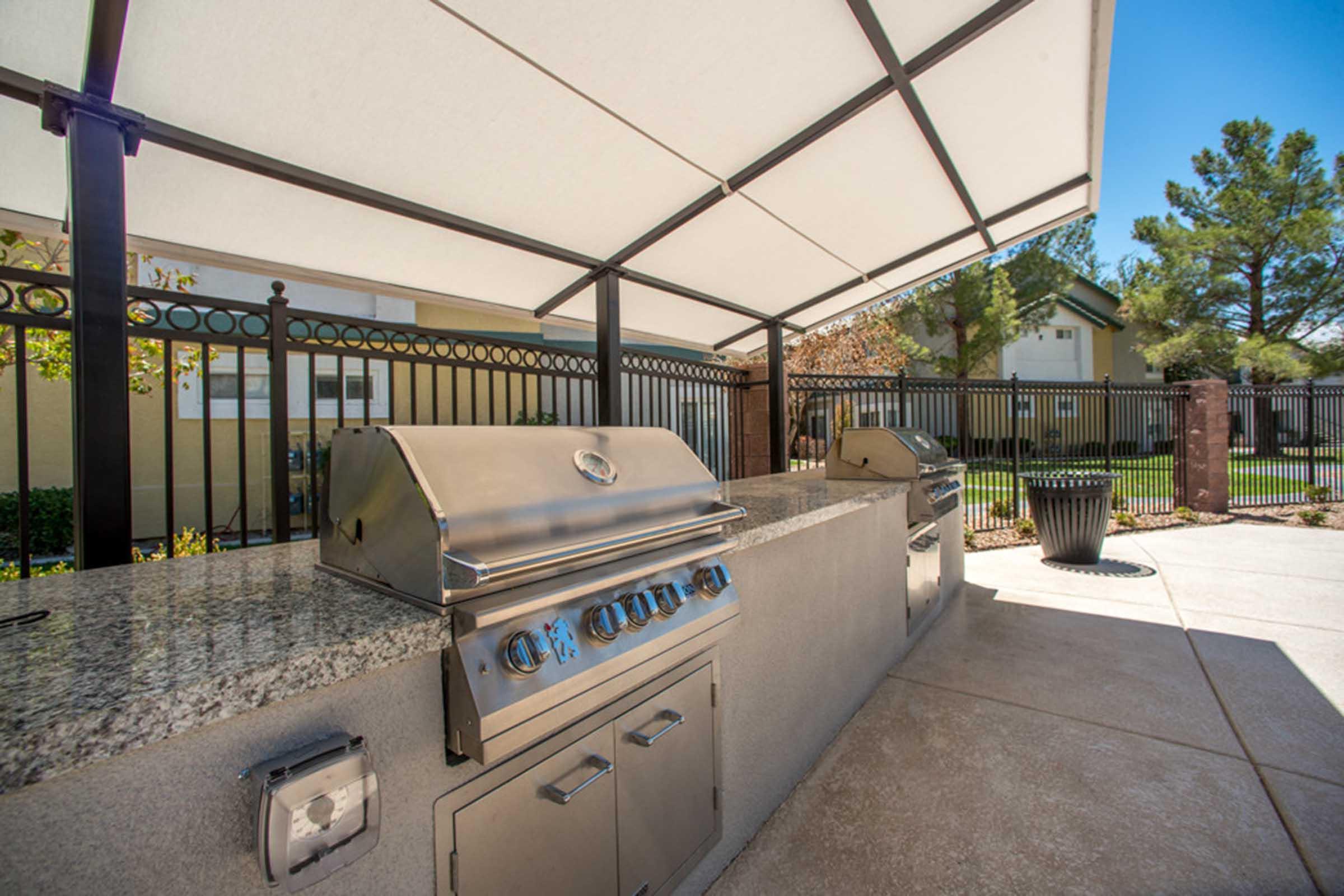 Outdoor grilling area featuring stainless steel gas grills under a shaded canopy. The space includes a granite countertop and is surrounded by a fence, with a view of a grassy area and buildings in the background. It's a sunny day with clear blue skies.