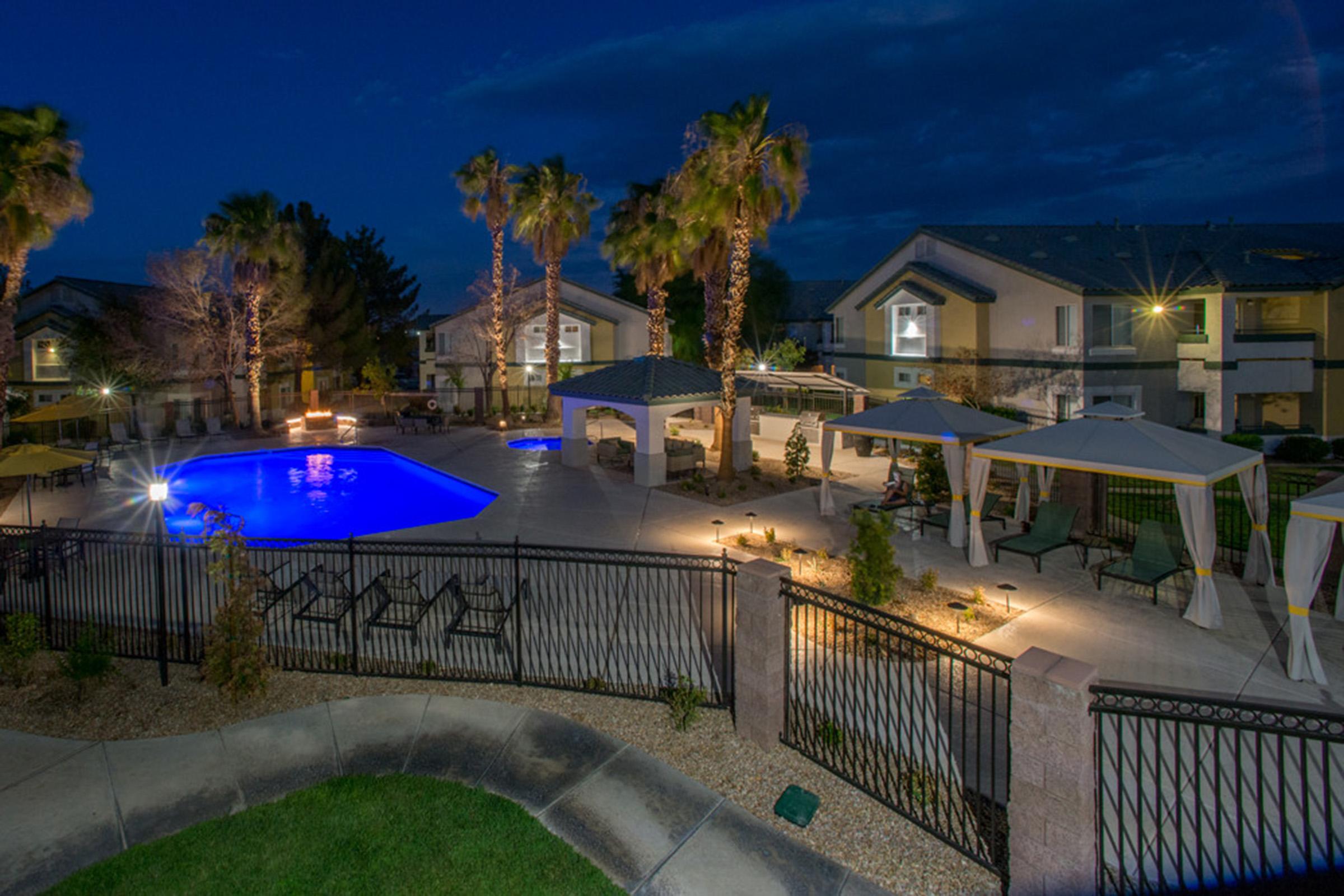 A beautifully landscaped pool area at night featuring a bright blue pool surrounded by lounge chairs, palm trees, and elegantly lit cabanas. The scene includes modern apartment buildings in the background, creating a serene and inviting atmosphere.
