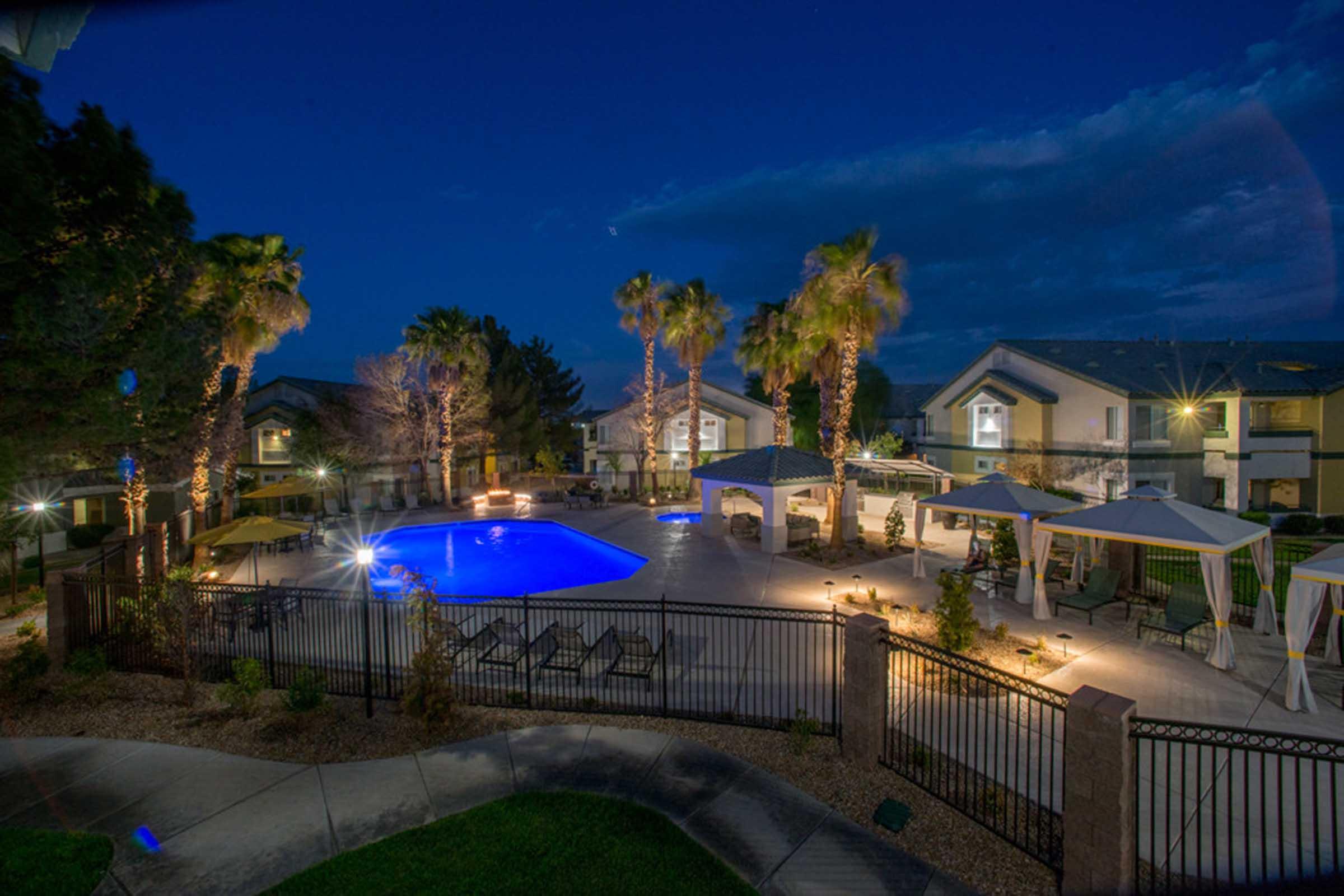 A well-lit outdoor pool area at night, featuring a large blue swimming pool surrounded by palm trees. There are lounge chairs and several cabanas around the pool. The atmosphere is inviting and serene, with soft lights illuminating the space and residential buildings in the background.